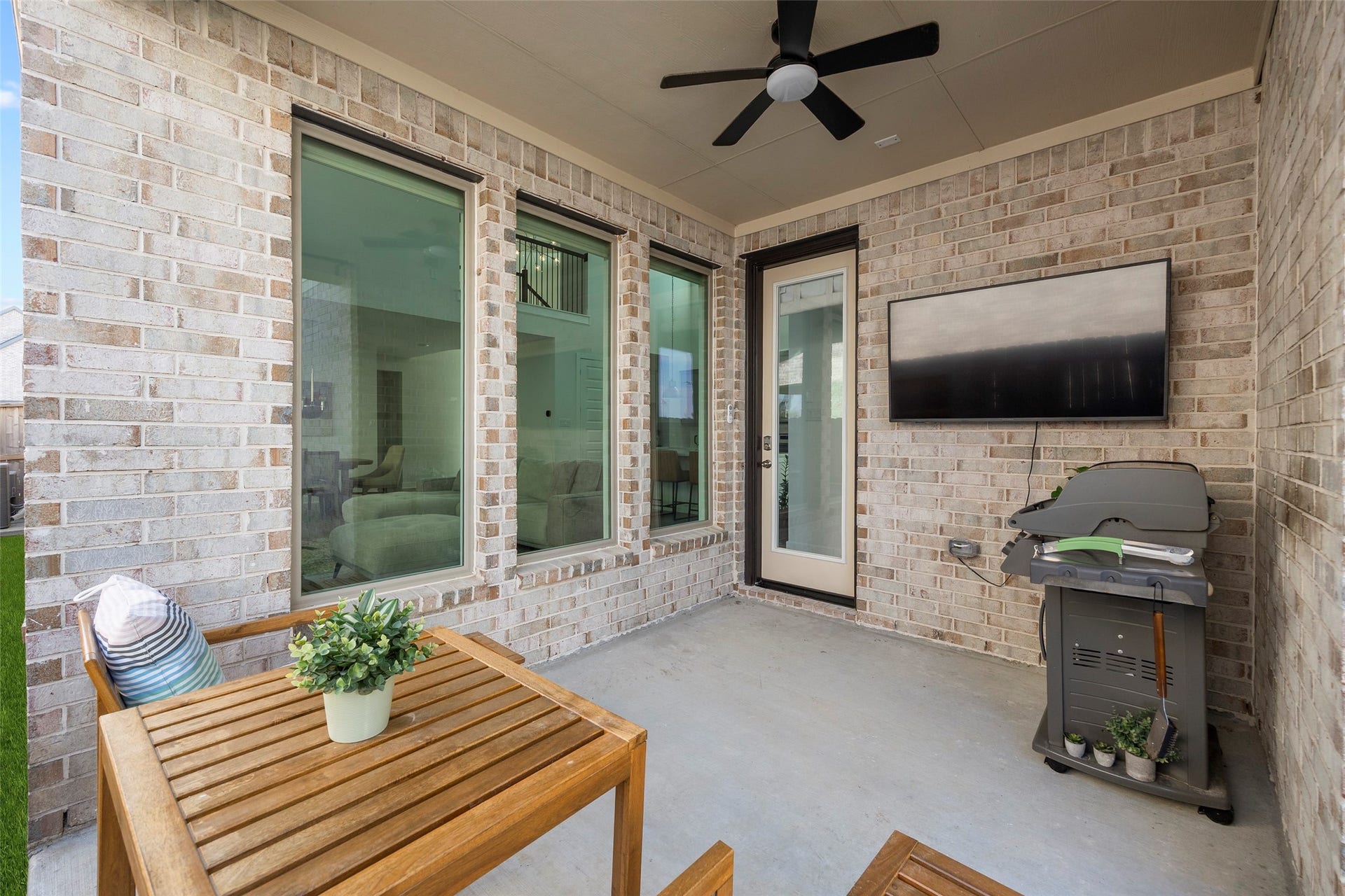 7311 Winding Aster Way, Katy, TX 77493 This reverse view of the covered patio shows the rear entry door, tall windows and brick backdrop. The sheltered space is currently arranged for outdoor dining and television viewing, illustrating its potential for relaxed evenings or weekend entertaining. A ceiling fan adds comfort above the seating area.
