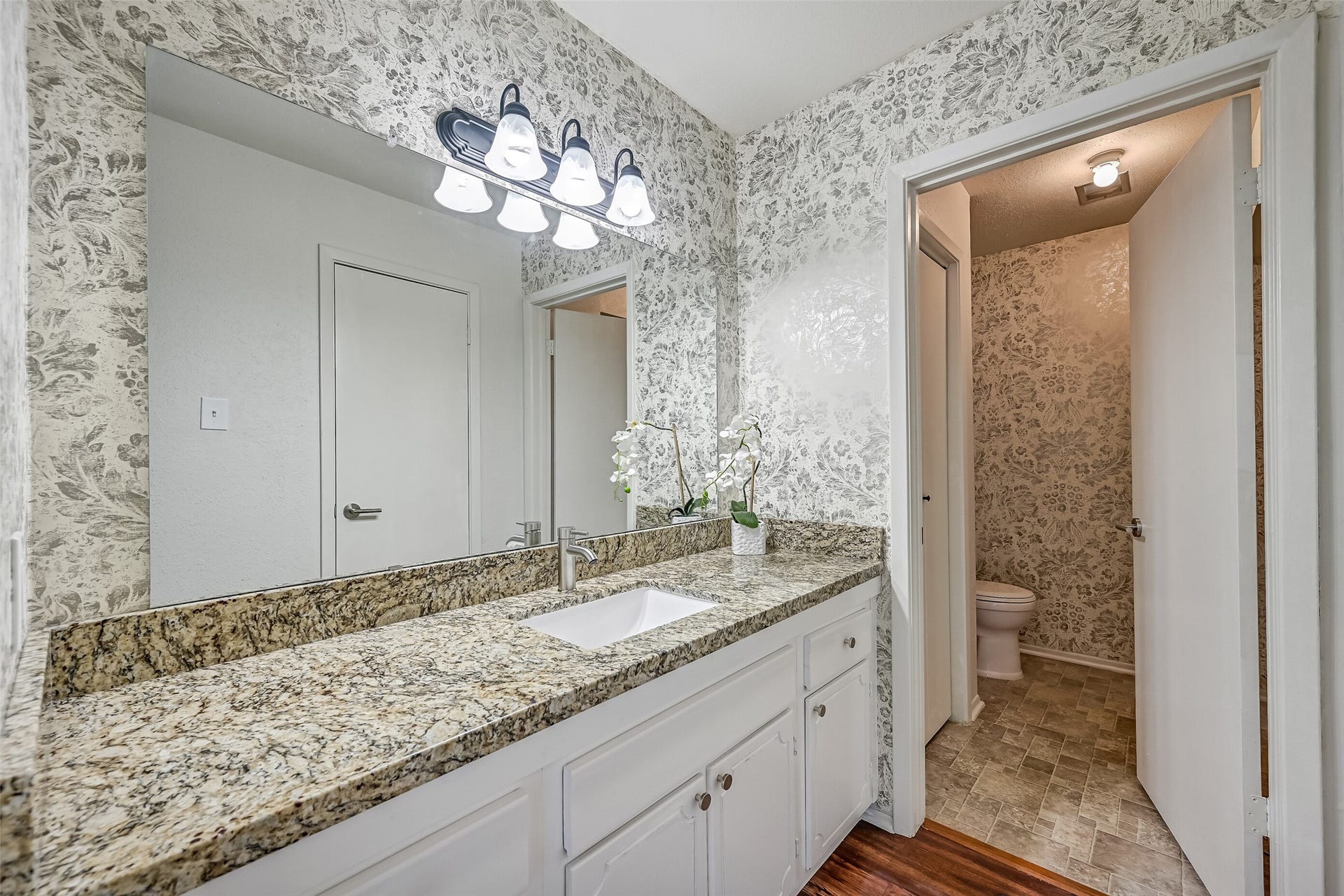 5005 Georgi Lane, #155, Houston, TX 77092 The primary bathroom features a separate vanity area with a granite countertop, undermount sink, updated faucet & contemporary vanity lighting. Decorative floral wallpaper adds character, while a large wall mirror & white cabinetry provide a functional space for getting ready each day.