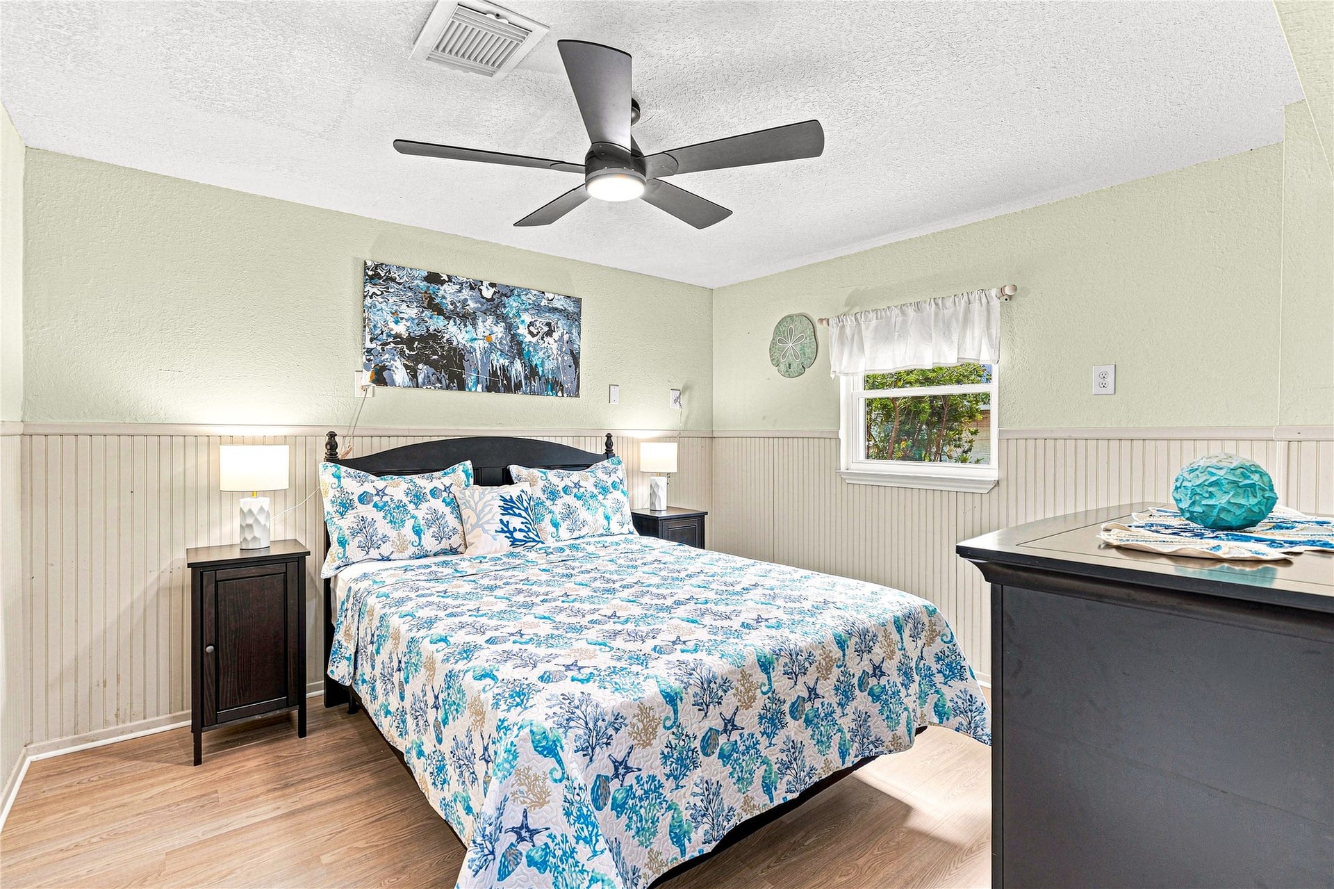 19 N Sandpiper Street, La Marque, TX 77568 Cozy bedroom with a modern ceiling fan, featuring a comfortable bed adorned with a vibrant blue and white quilt. Natural light streams through a window, complementing the soft, neutral wall tones and wainscoting.