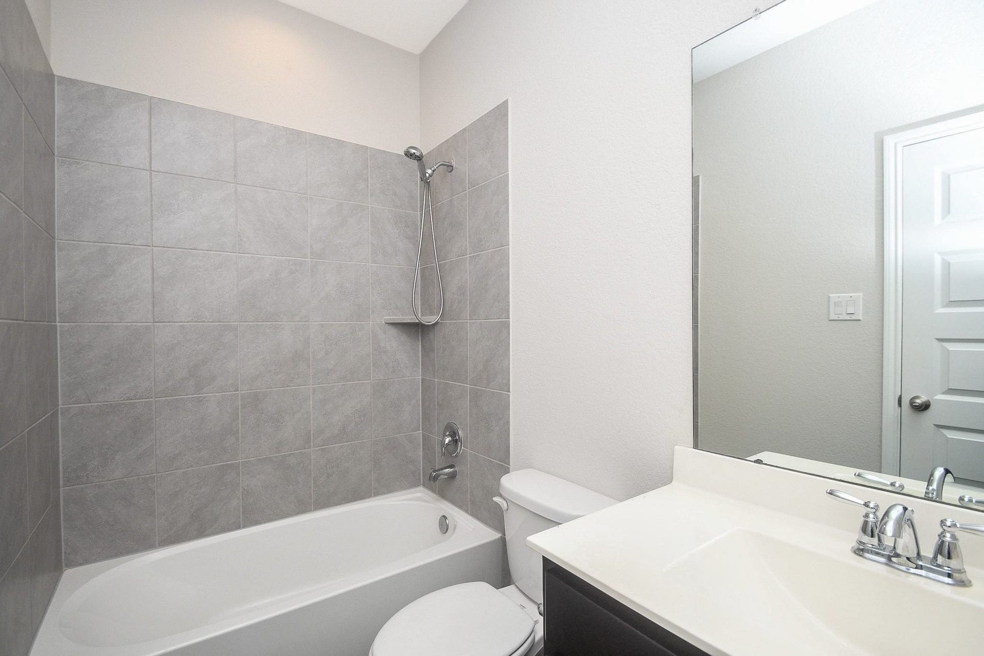 Modern bathroom downstairs featuring sleek gray tile, a pristine bathtub with shower, and a spacious vanity with a large mirror for a clean and contemporary look next to the secondary bedroom.