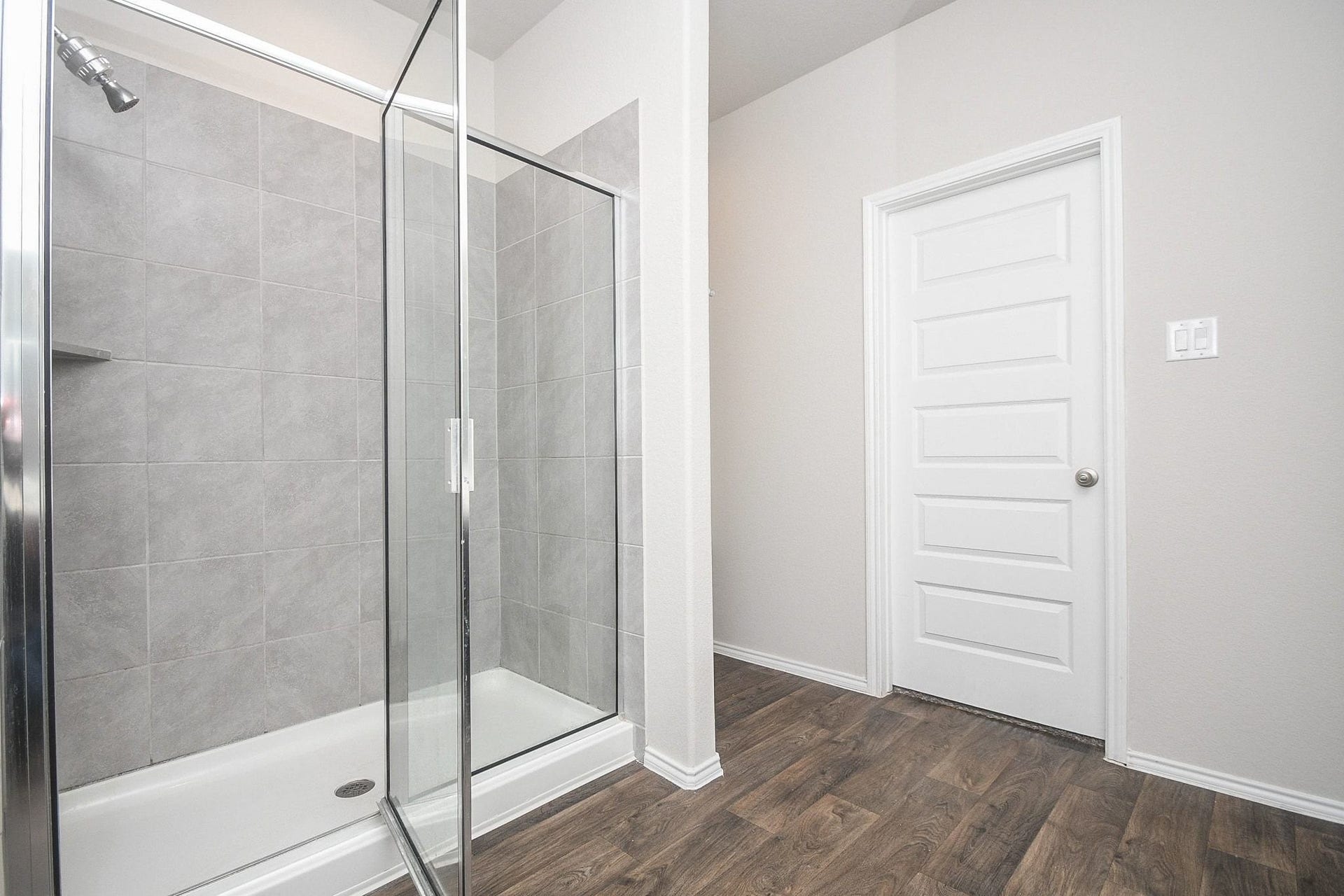 Modern primary bathroom featuring a sleek glass-enclosed shower with elegant gray tile work, complemented by warm wood-style flooring and a crisp white door.