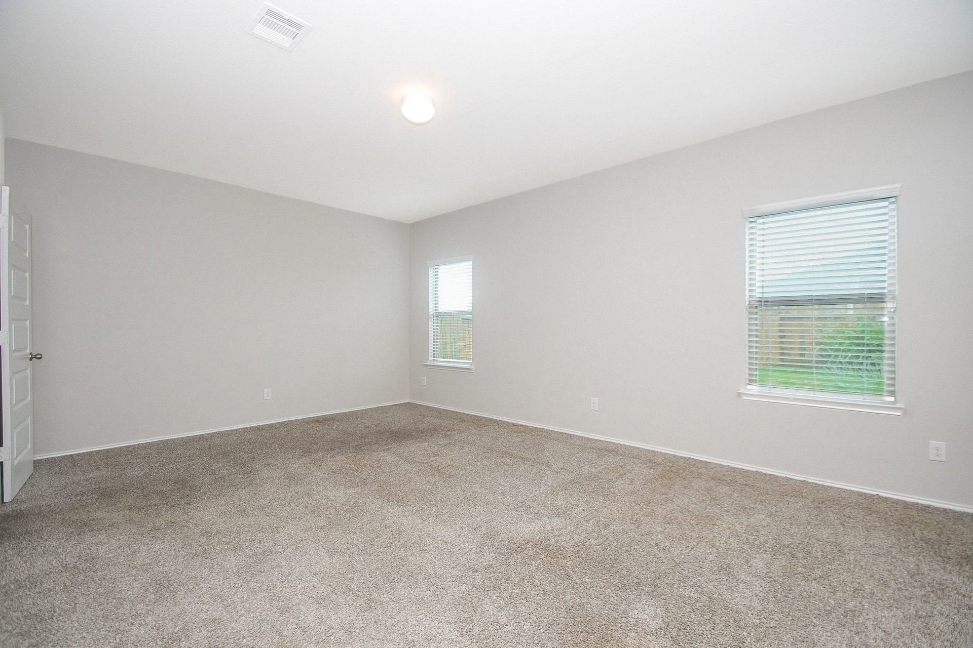 Spacious, light-filled primary bedroom with neutral walls and plush carpeting, featuring two windows that offer a view of the outdoors.