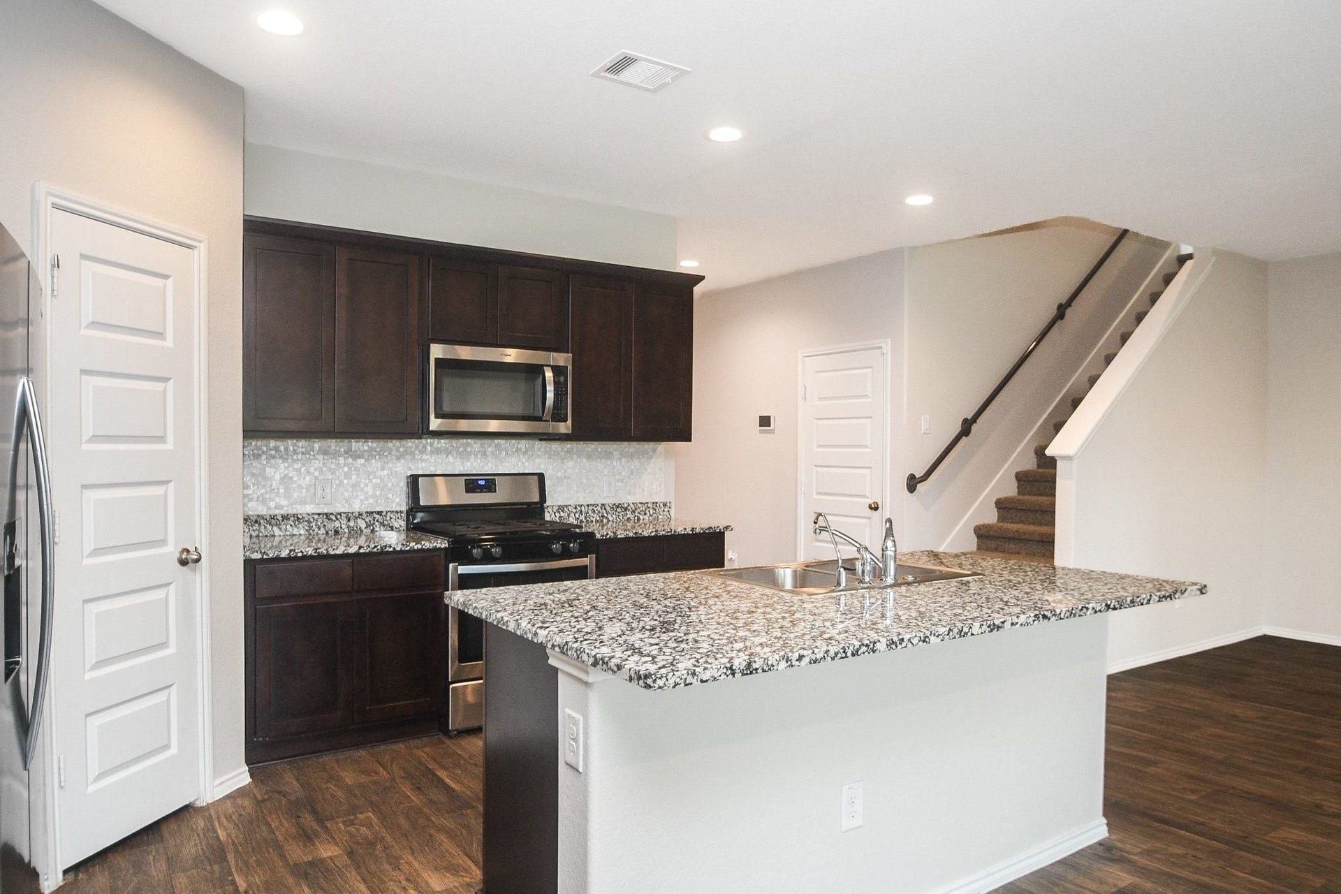 View of the modern kitchen with dark wood cabinets, granite countertops, and stainless steel appliances, adjacent to an open staircase.