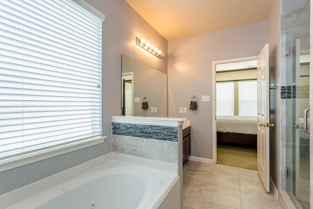 4530 Hall Croft Chase Lane, Katy, TX 77449 Experience relaxation in this elegant bathroom featuring a spacious soaking tub, modern vanity with stylish tile accents, and a glass-enclosed shower. Natural light filters through the large window, enhancing the serene ambiance.