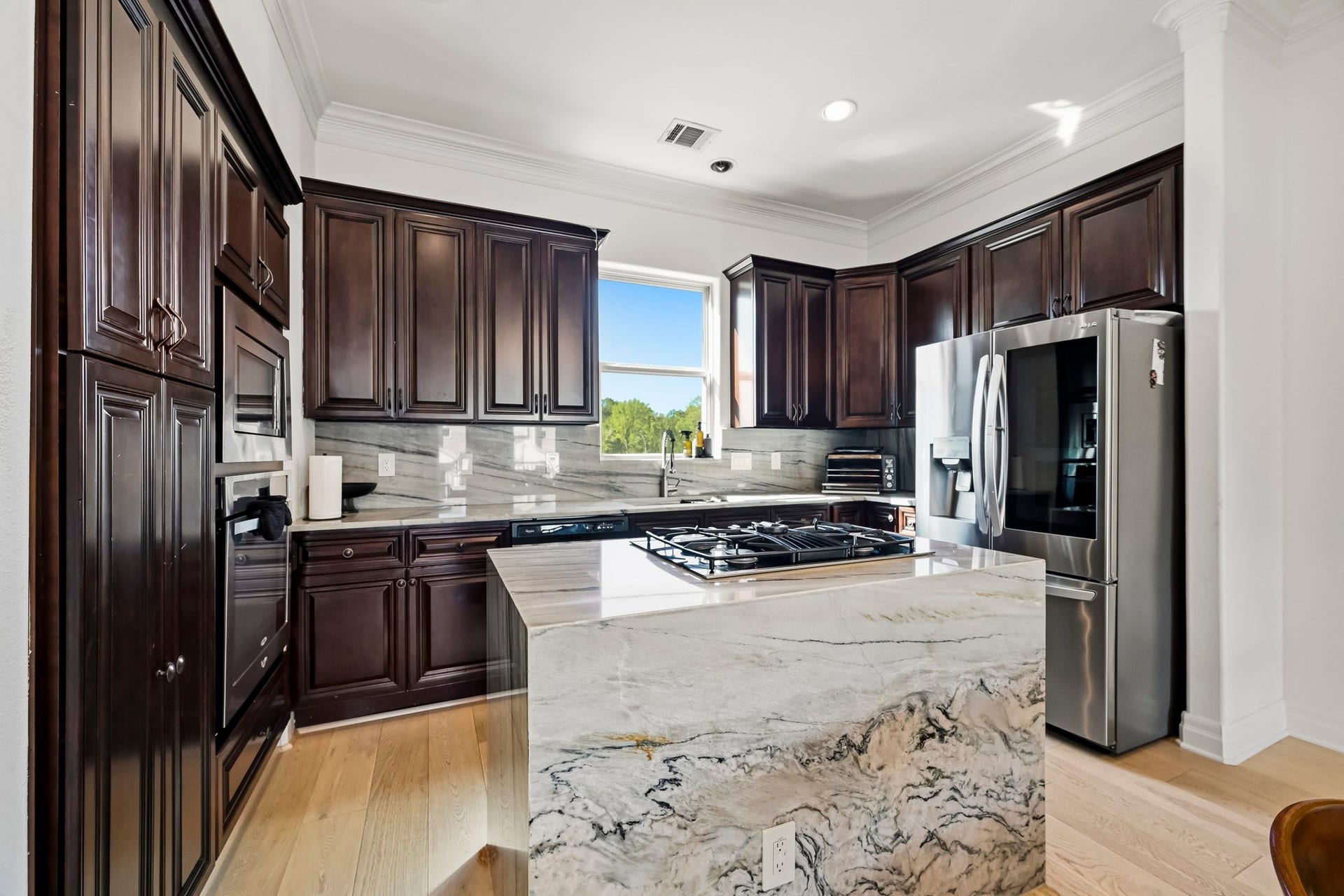 11009 Upland Forest Drive, Houston, TX 77043 Elegant kitchen featuring rich dark wood cabinetry, a sleek stainless steel refrigerator, and a striking marble island with a built-in gas cooktop, all bathed in natural light from a well-placed window.