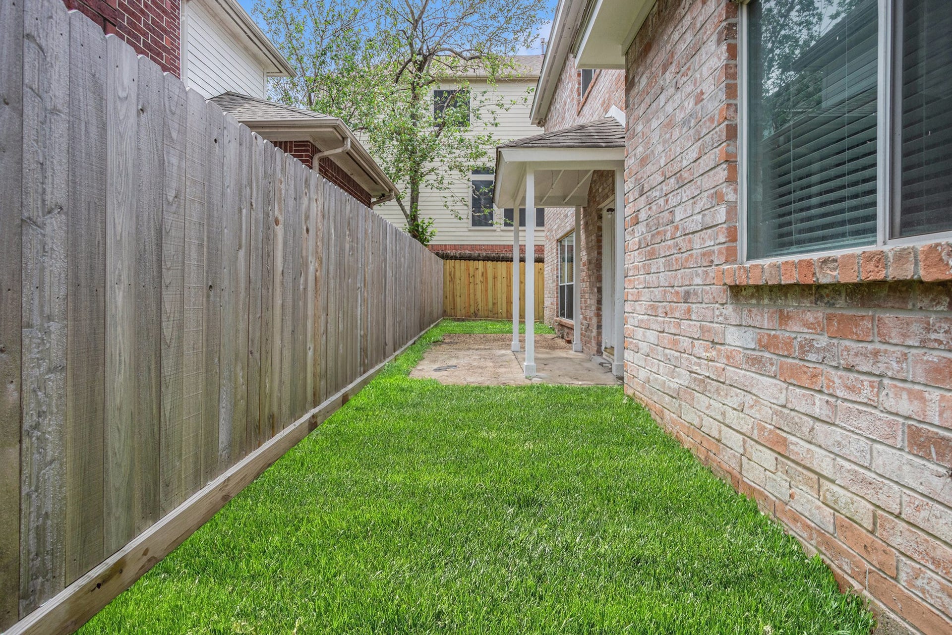 5001 Jackson Street, Houston, TX 77004 This photo shows a fenced backyard with a well-maintained grassy area, bordered by a wooden fence and a brick exterior wall of the house. There is a small covered patio area with a concrete path leading to a wooden gate.