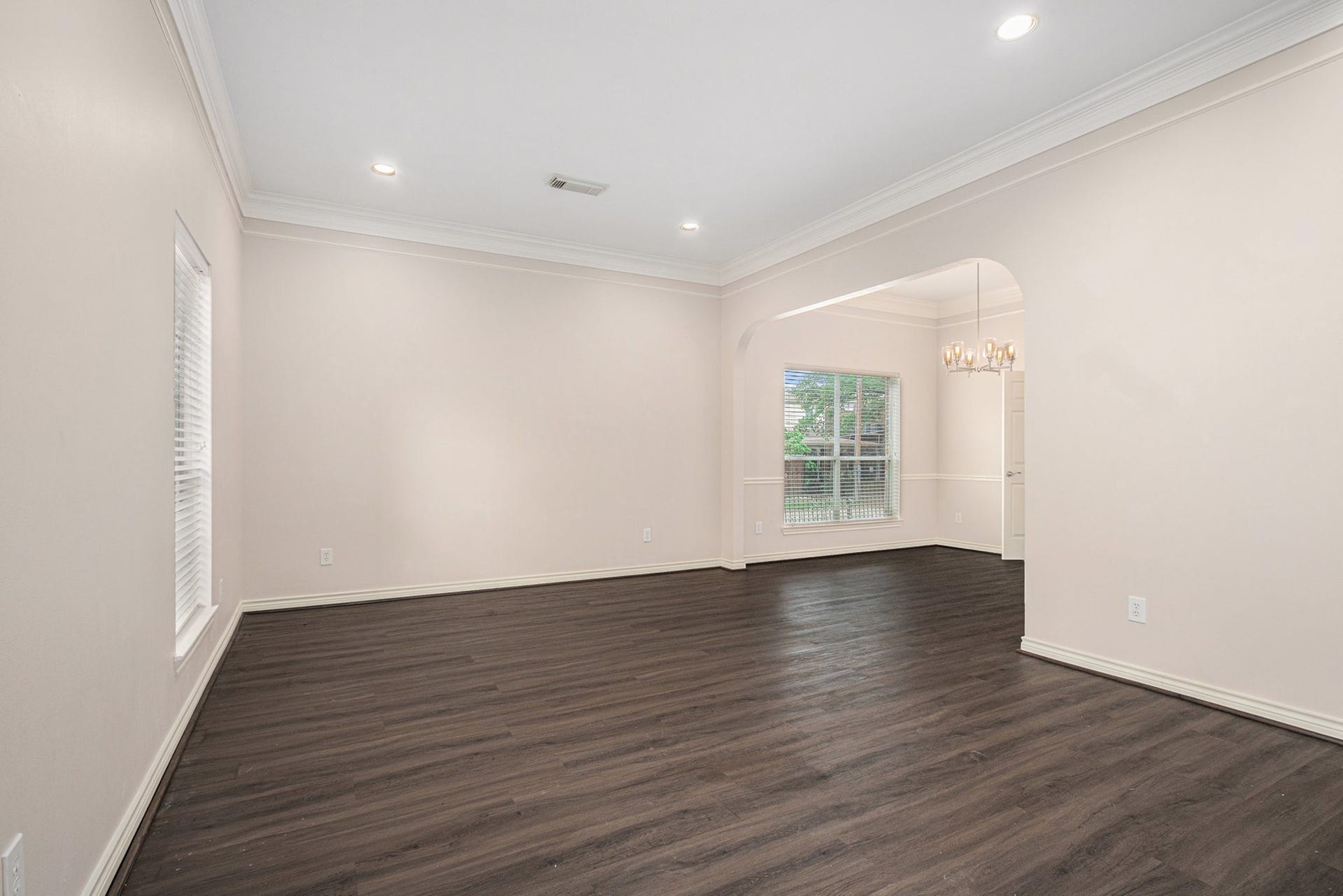 5001 Jackson Street, Houston, TX 77004 This spacious room features dark wood flooring, soft cream walls, and recessed lighting. It includes a large window for natural light and an arched opening leading to an adjacent area with a chandelier.