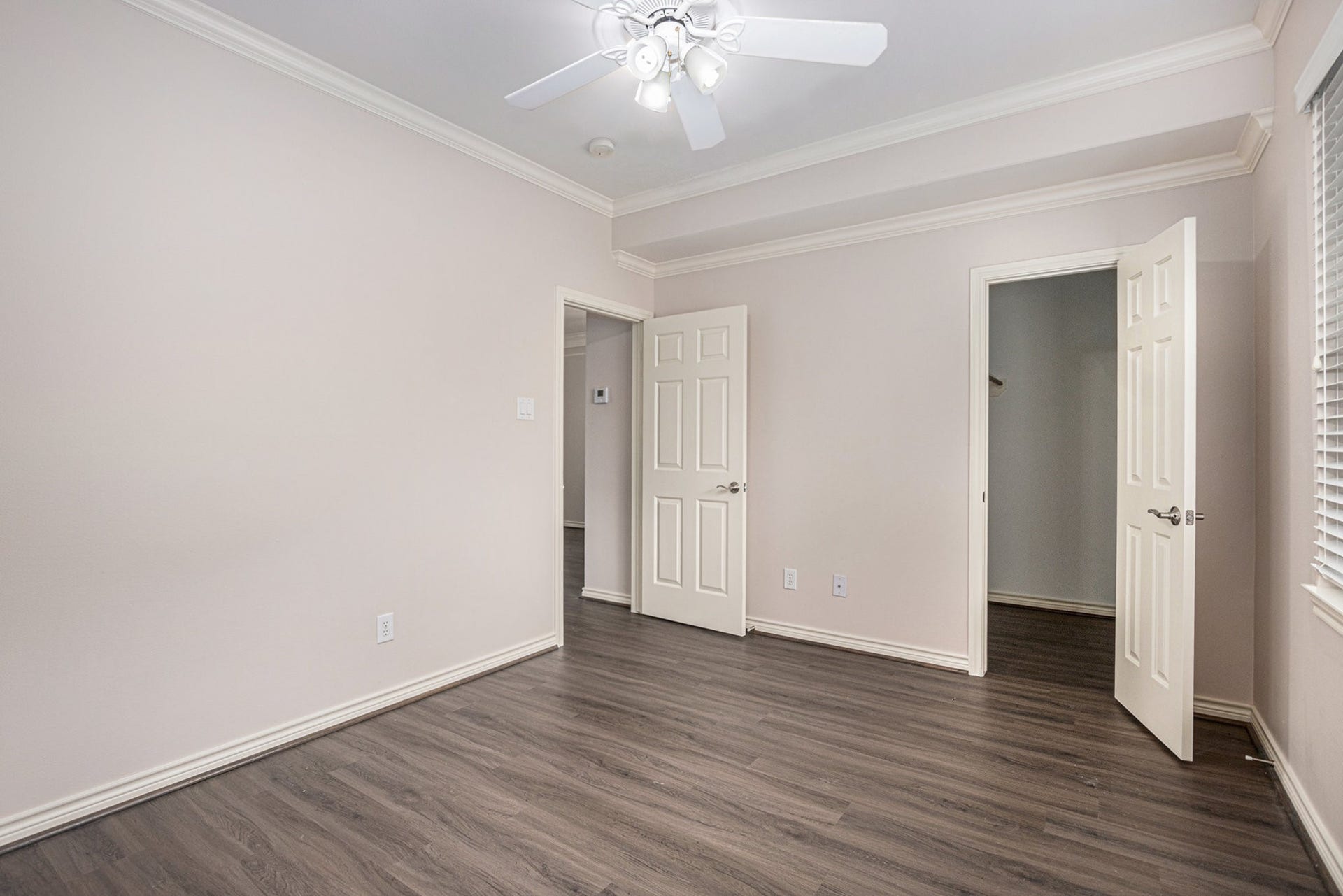 5001 Jackson Street, Houston, TX 77004 This photo shows a bright, empty room with light-colored walls and dark hardwood floors. It features a ceiling fan, crown molding, and two doorsone leading to a closet and the other to a hallway. There's a window on the right, allowing natural light to enter.