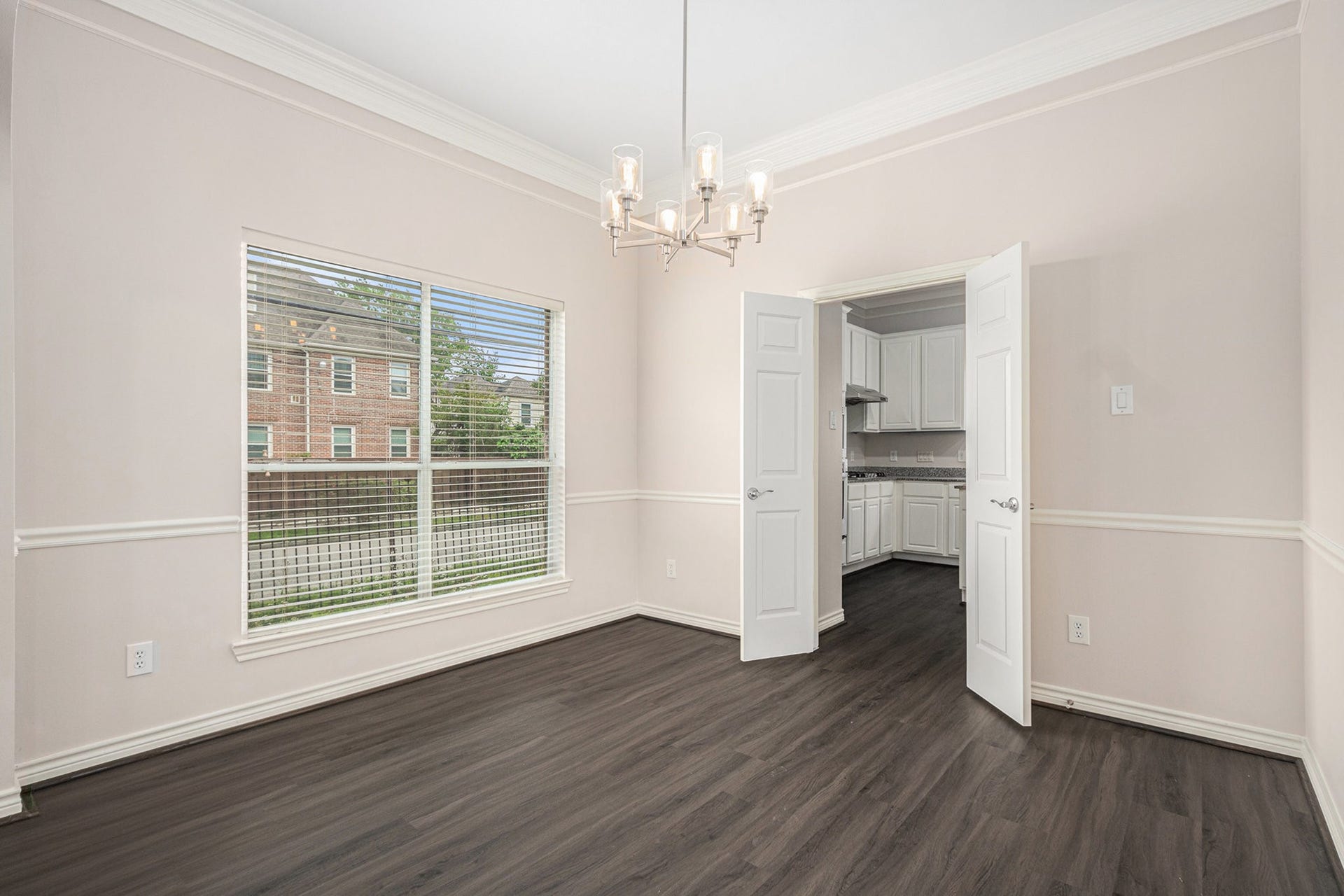 5001 Jackson Street, Houston, TX 77004 This photo shows a bright dining room with elegant crown molding, a modern chandelier, and dark wood flooring. Large windows provide natural light, and double doors lead into a white kitchen.