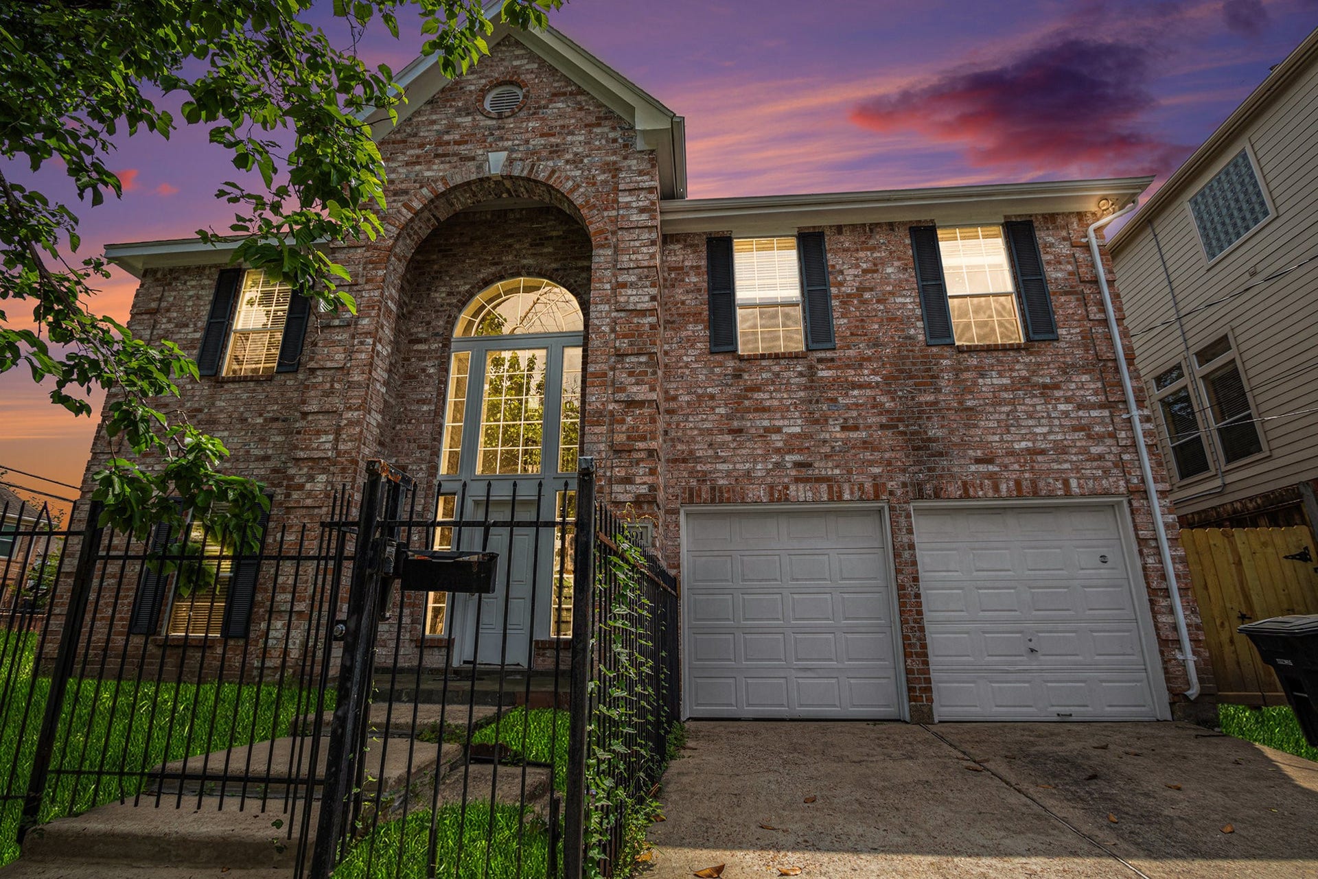 5001 Jackson Street, Houston, TX 77004 This photo features a two-story brick home with an elegant arched entryway and large windows. It has a double garage and is surrounded by a neatly fenced yard. The sunset sky adds a warm, inviting glow to the property.