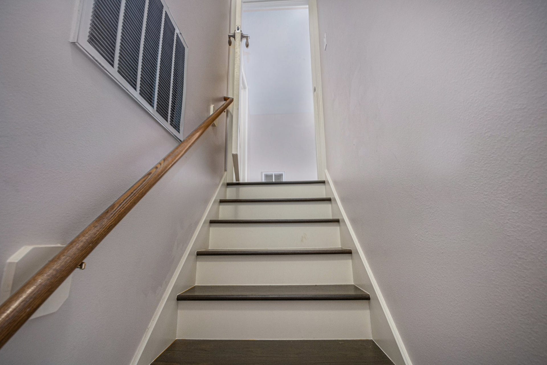5001 Jackson Street, Houston, TX 77004 This photo shows a staircase with dark wood steps and white risers, leading up to a doorway. The walls are painted a light neutral color, and a wooden handrail lines the left side. An air vent is visible on the left wall.