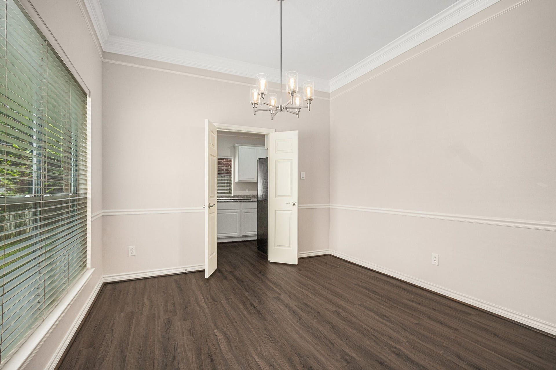 5001 Jackson Street, Houston, TX 77004 This room features elegant wood flooring, a large window with blinds for natural light, and a modern chandelier. It opens into a kitchen area through double doors, offering a seamless flow between spaces. Perfect for dining or as a versatile living area.
