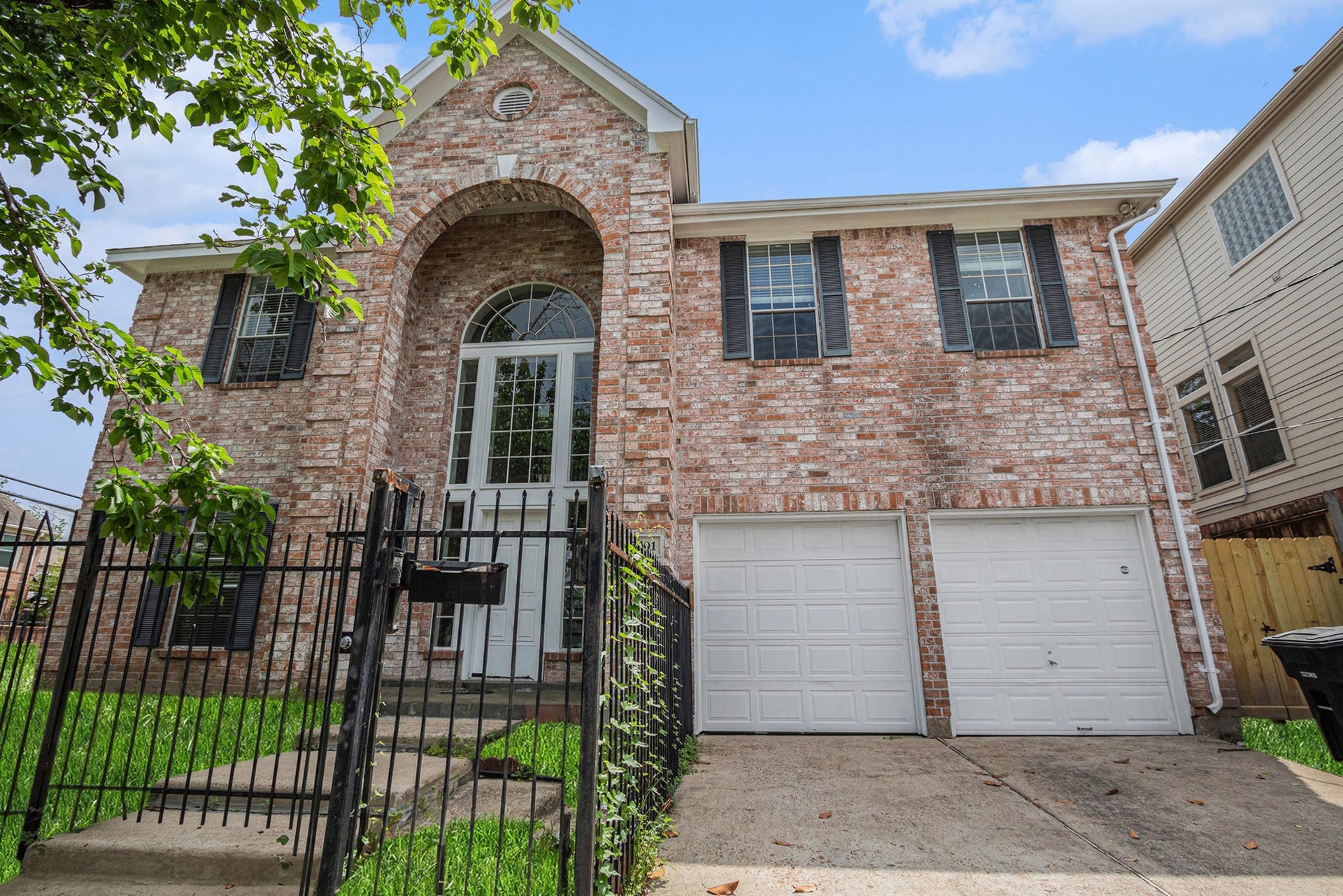 5001 Jackson Street, Houston, TX 77004 This photo showcases a two-story brick home with an arched entryway and large windows. It features a fenced front yard, a double garage, and a concrete driveway, offering a welcoming and secure exterior.
