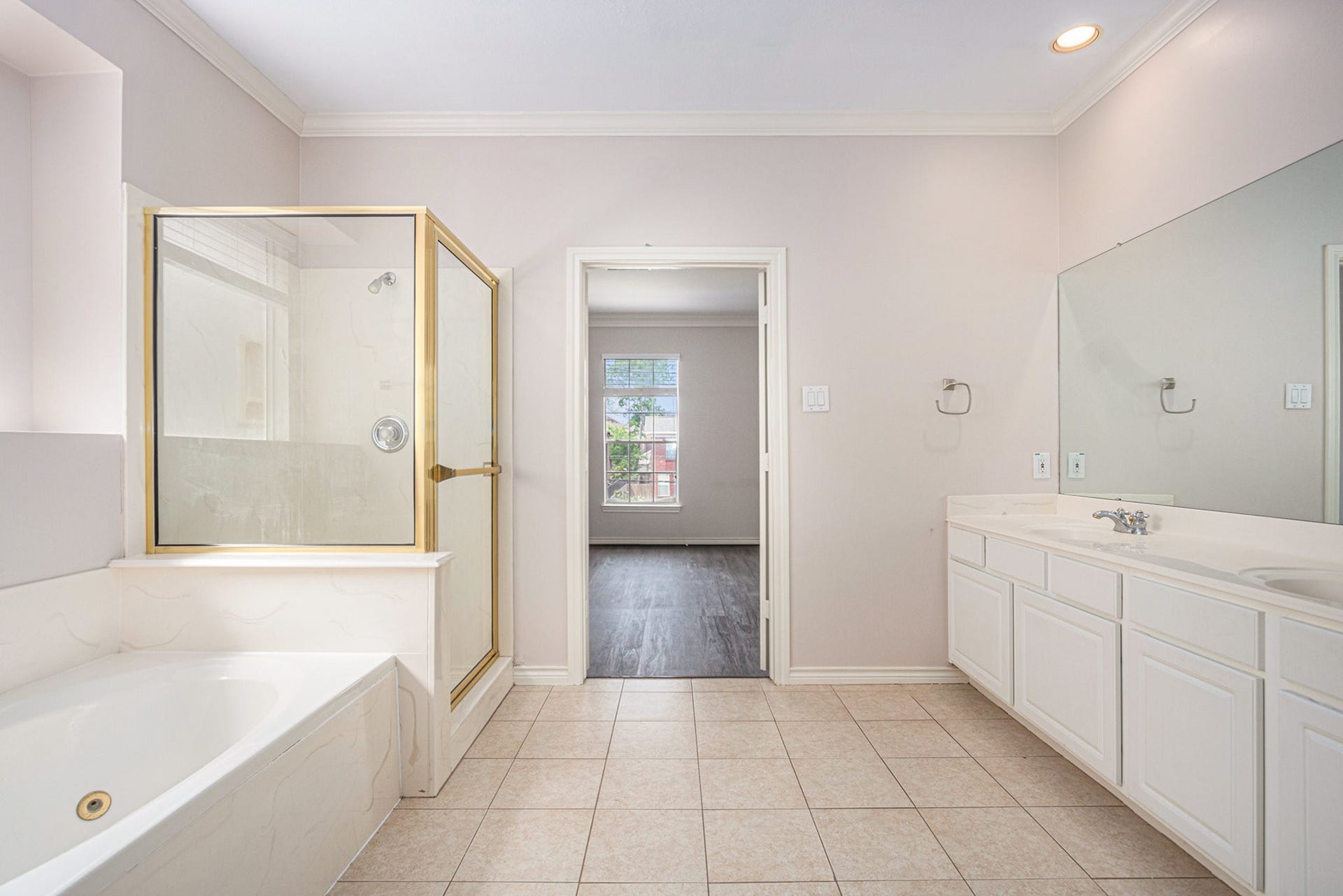 5001 Jackson Street, Houston, TX 77004 This photo showcases a spacious bathroom with a large soaking tub, glass-enclosed shower, dual-sink vanity, and neutral tile flooring. A doorway leads to an adjacent room with a window.
