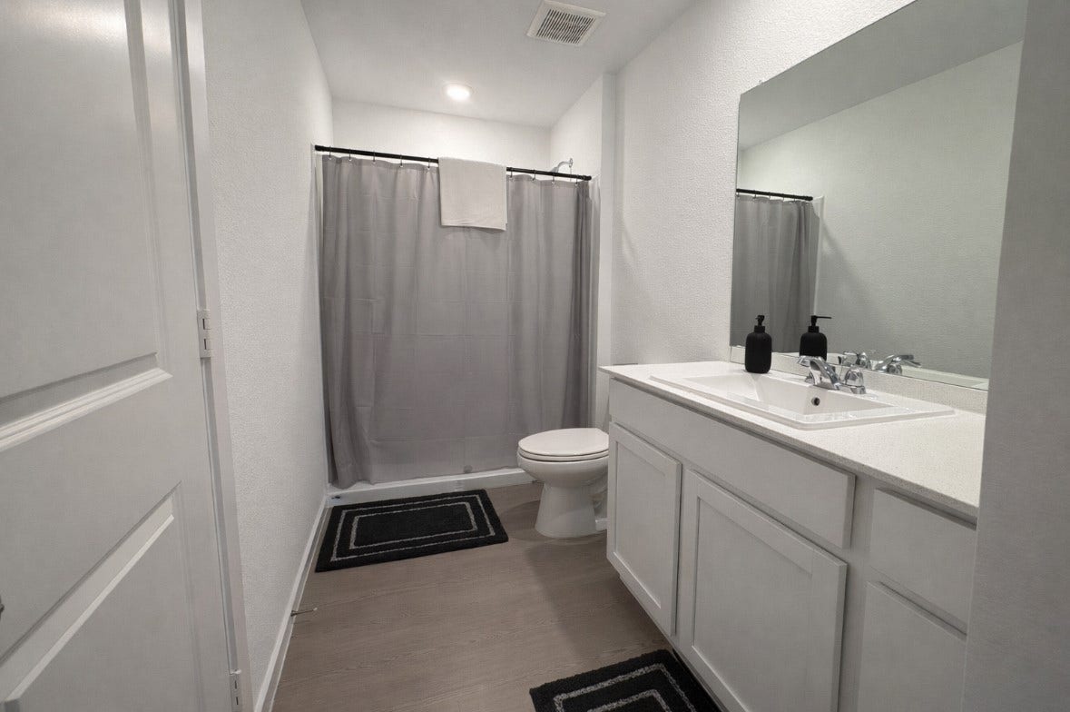 21151 Vercelli, New Caney, TX 77357 This bathroom features a clean, modern design with neutral colors. It includes a large mirror, a vanity with ample storage, and a shower area with a curtain. The flooring complements the minimalist aesthetic, making it a practical and stylish space.