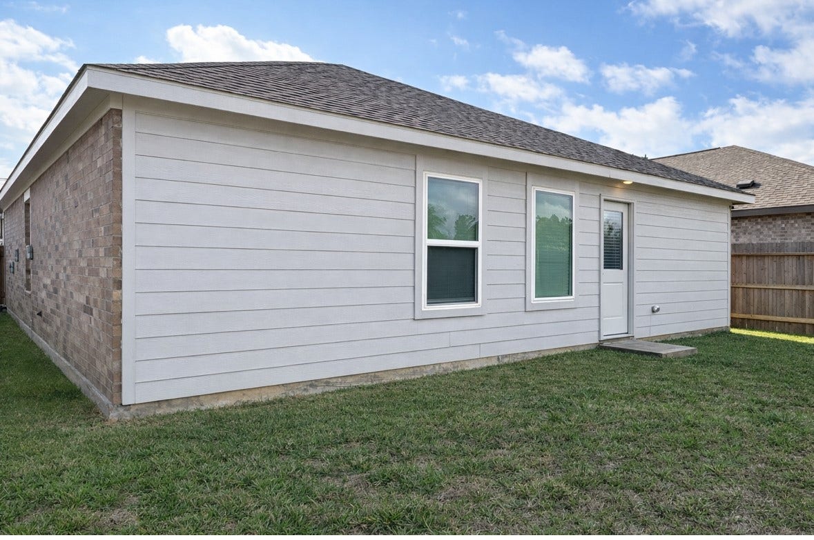 21151 Vercelli, New Caney, TX 77357 This photo shows the back exterior of a single-story home with a combination of brick and siding. It features two windows and a door leading to a grassy backyard, enclosed by a wooden fence. A small concrete step is present at the door.