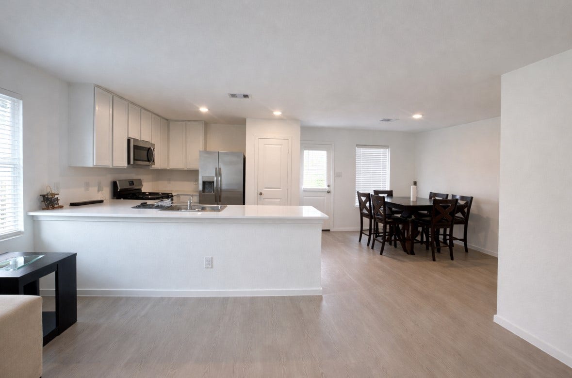 21151 Vercelli, New Caney, TX 77357 This photo showcases a modern, open-concept kitchen and dining area. The kitchen features sleek white cabinetry, stainless steel appliances, and a large countertop. The dining space includes a stylish table with seating for six, and the room has ample natural light from multiple windows.