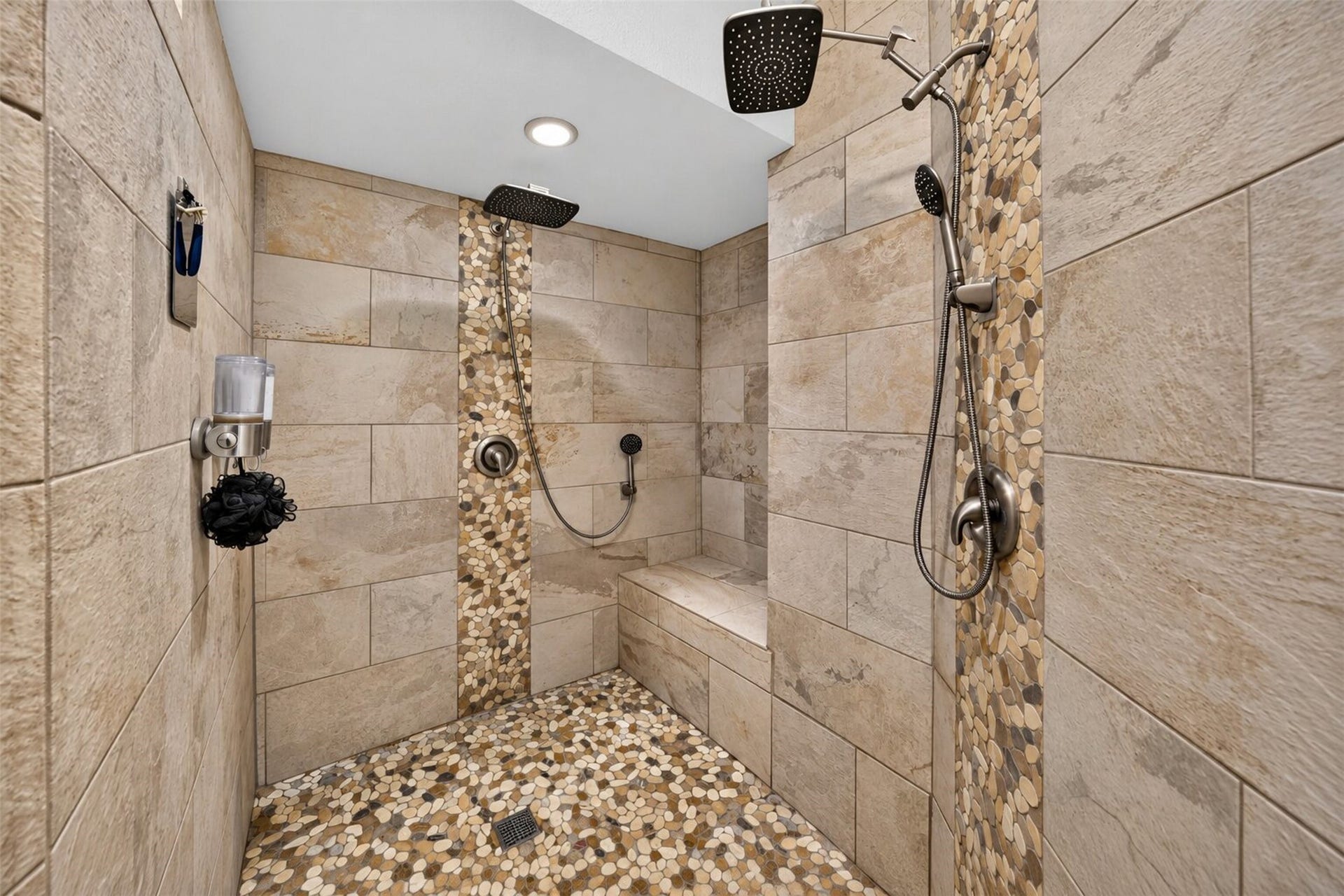 Walk-in shower features two overhead showerheads, handheld sprayers, a built-in bench, and pebble-pattern flooring.