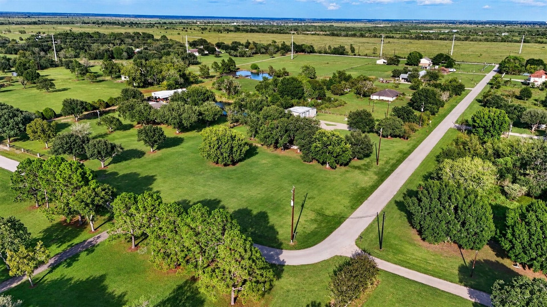 Wide aerial view shows the property's setting among open fields, mature trees, and nearby homes.