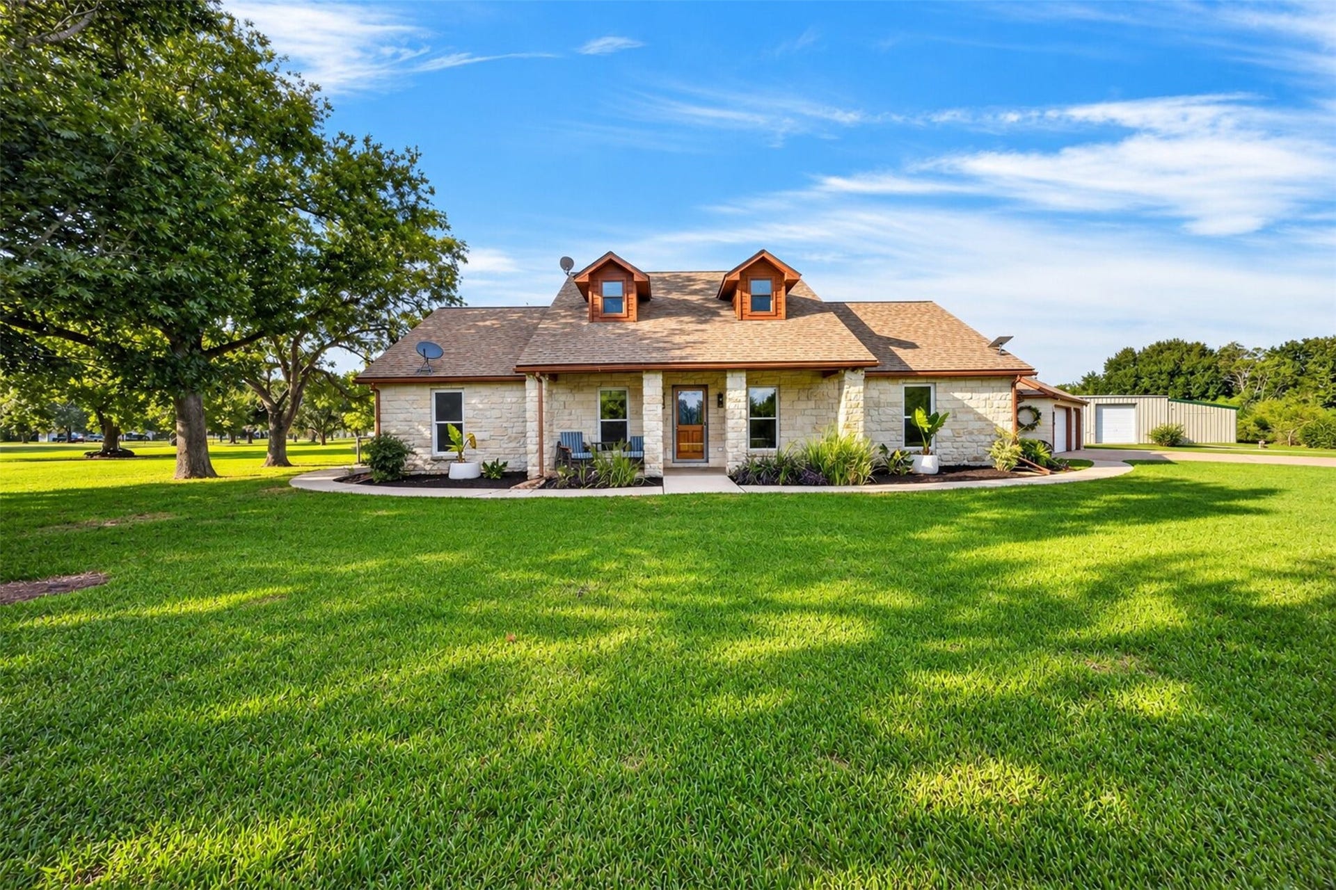Stone-front home with twin dormers, a covered entry porch, landscaped beds, and mature shade trees.