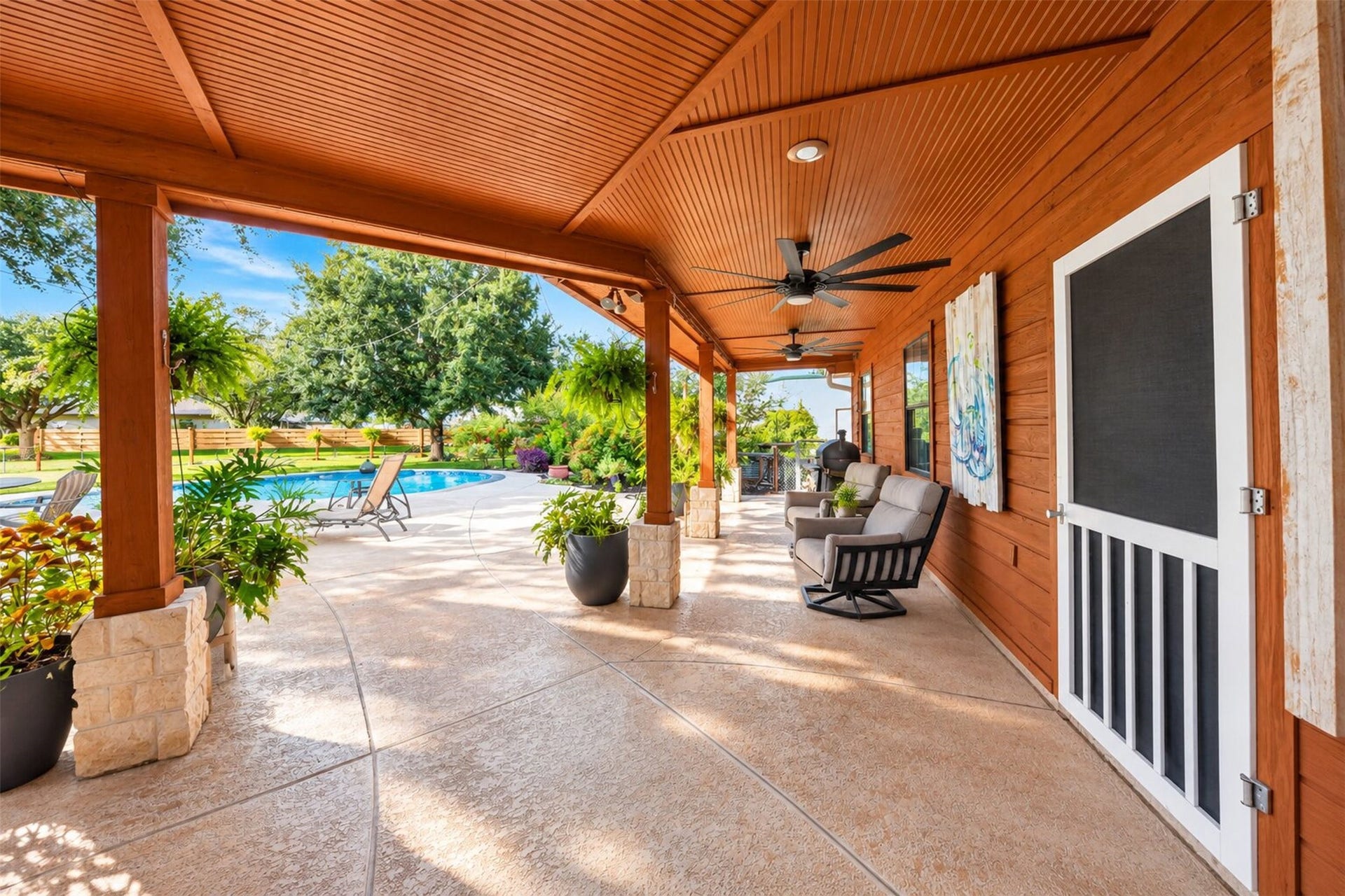 Covered rear porch features a wood ceiling, ceiling fans, stone-accented columns, and pool views.