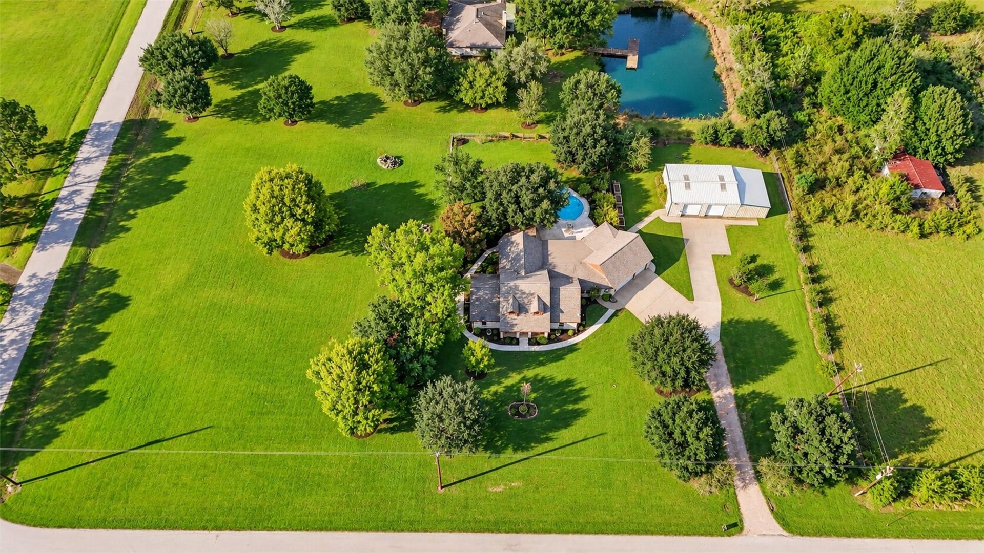 Aerial overview shows the home, pool, detached metal building, driveway, and surrounding open lawn.