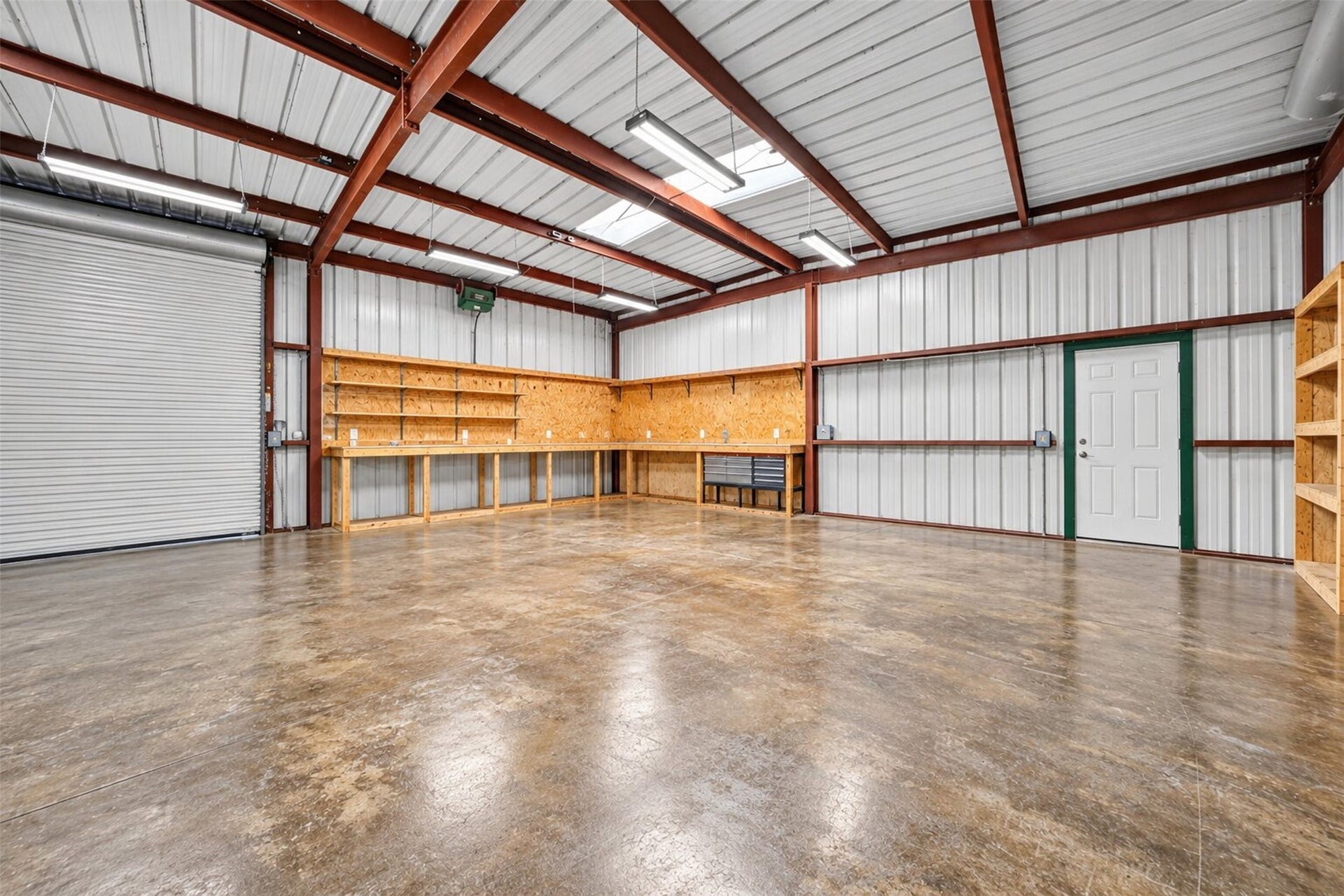 Metal workshop interior features a long workbench, wall shelving, overhead lighting, and a roll-up door.
