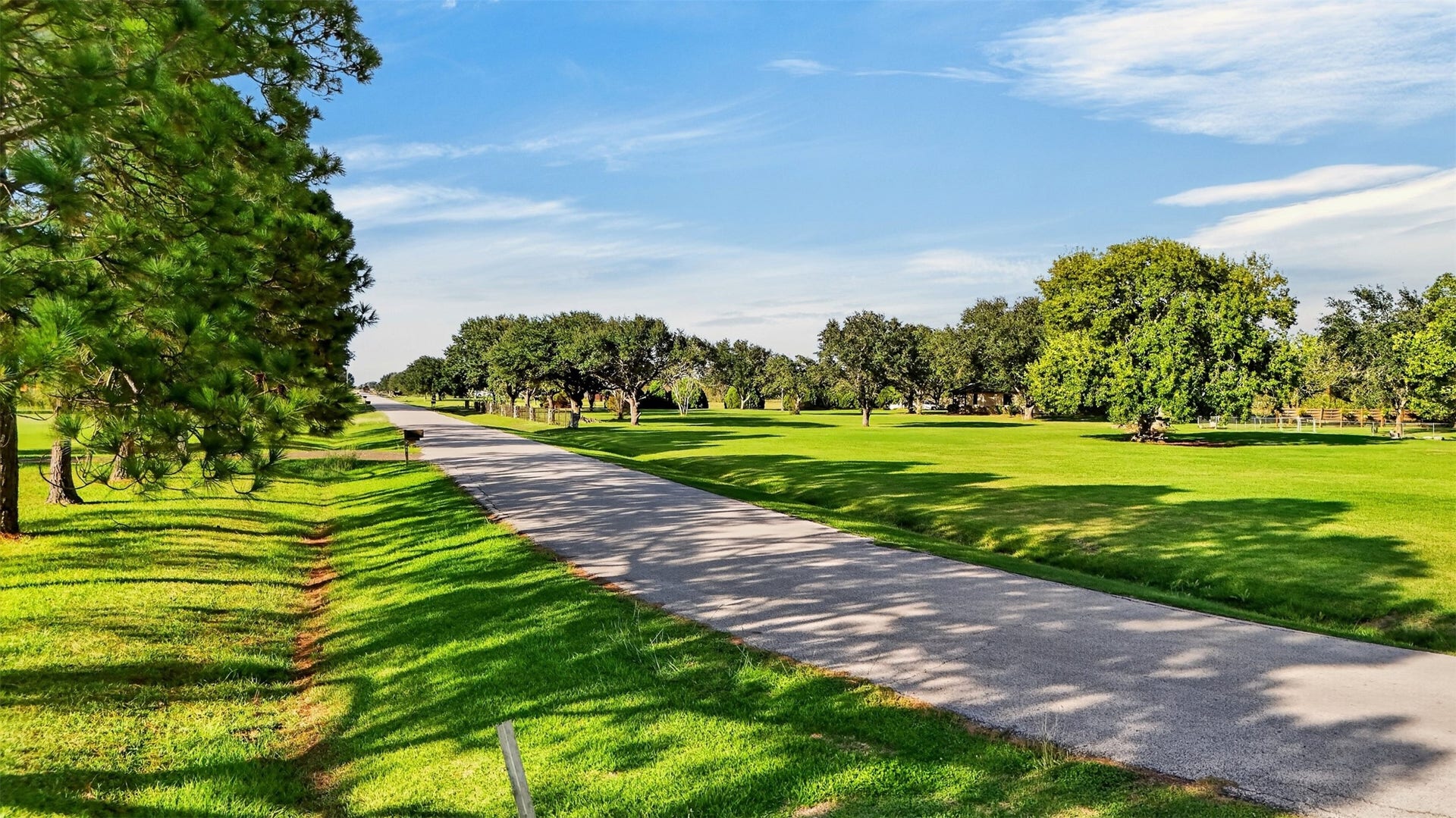 View along the paved road shows grassy frontage, mature trees, and neighboring rural homes.