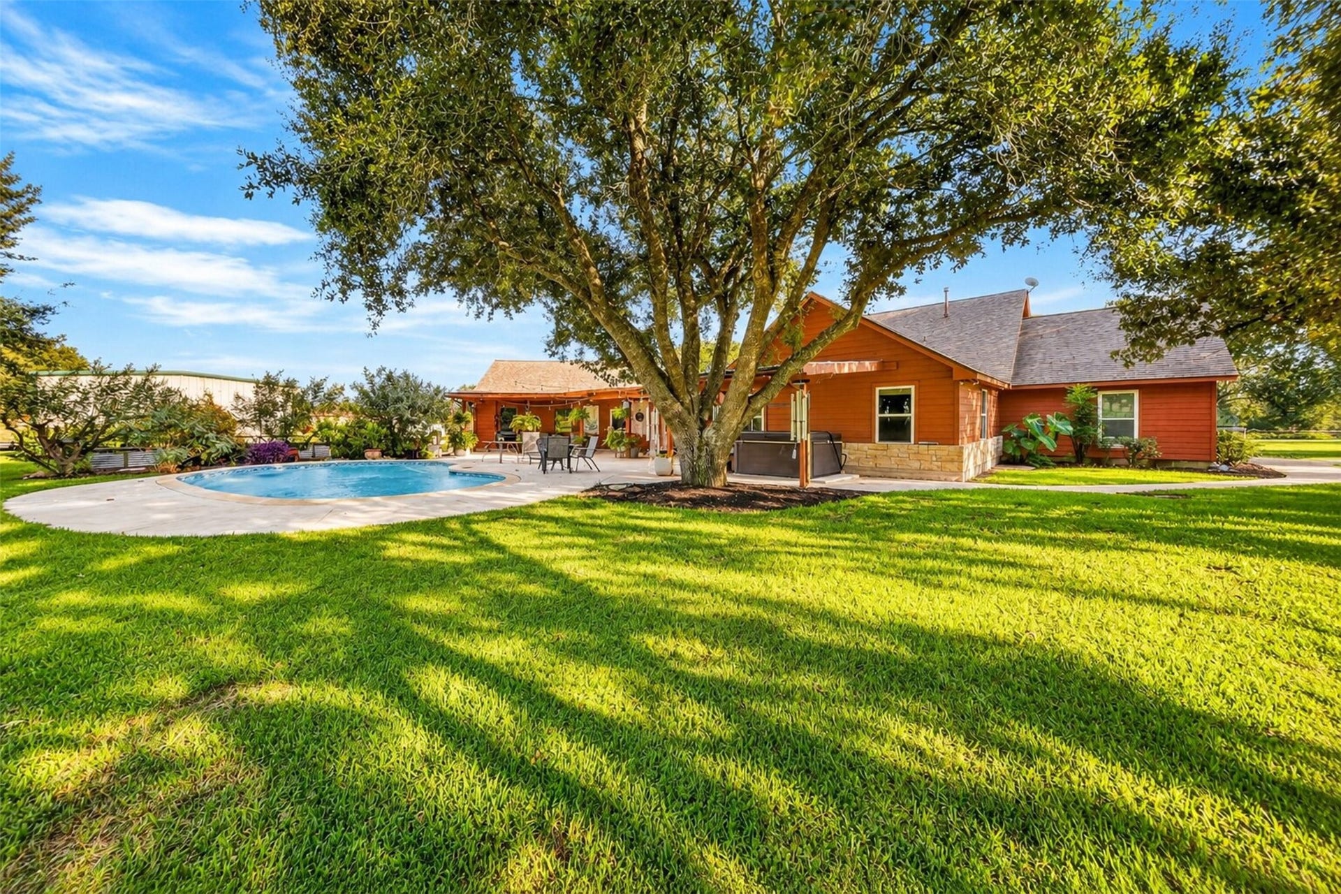 Rear exterior overlooks the pool, expansive patio, and lawn shaded by mature trees.