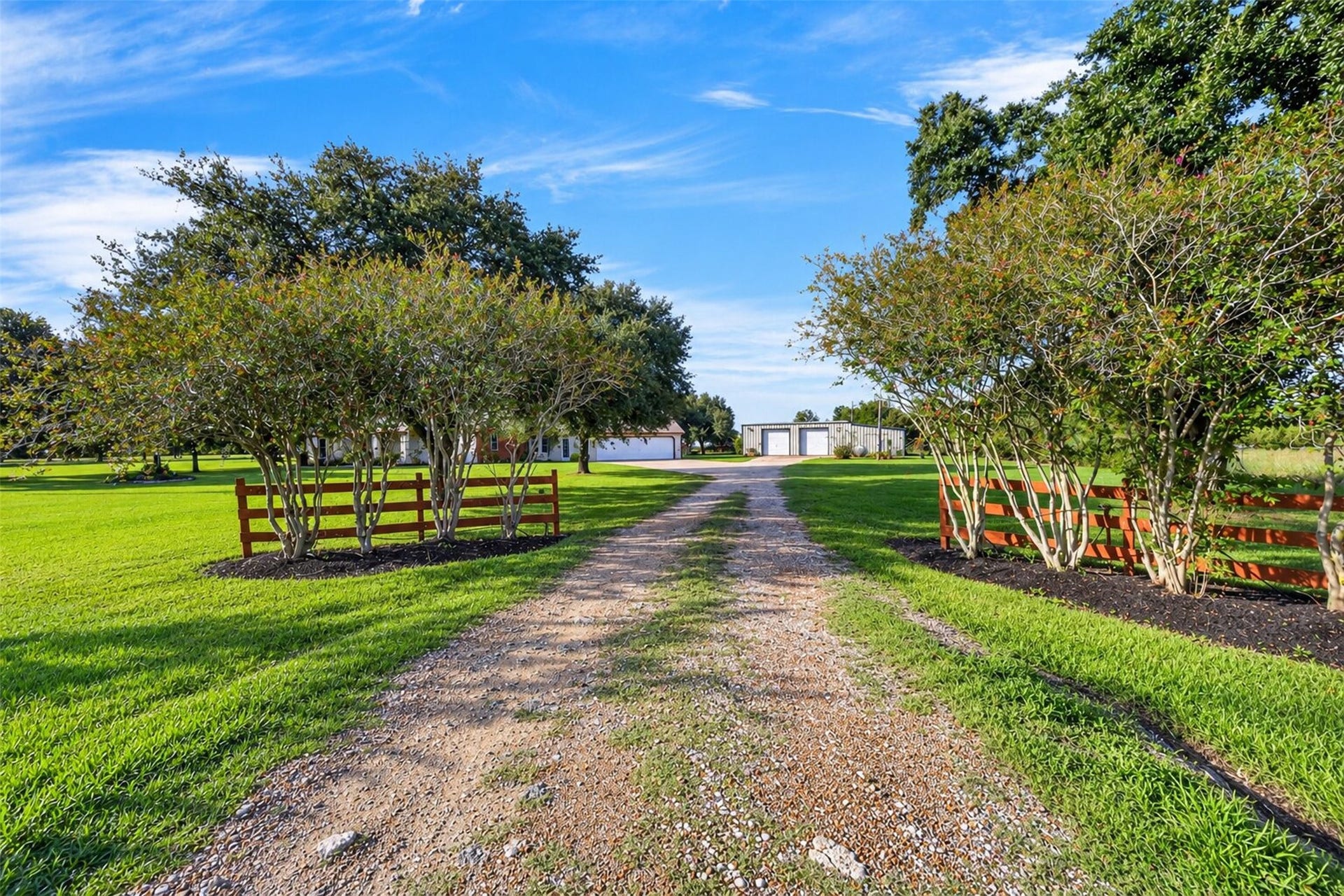 Gravel driveway entrance framed by landscaped trees and sections of wood rail fencing.