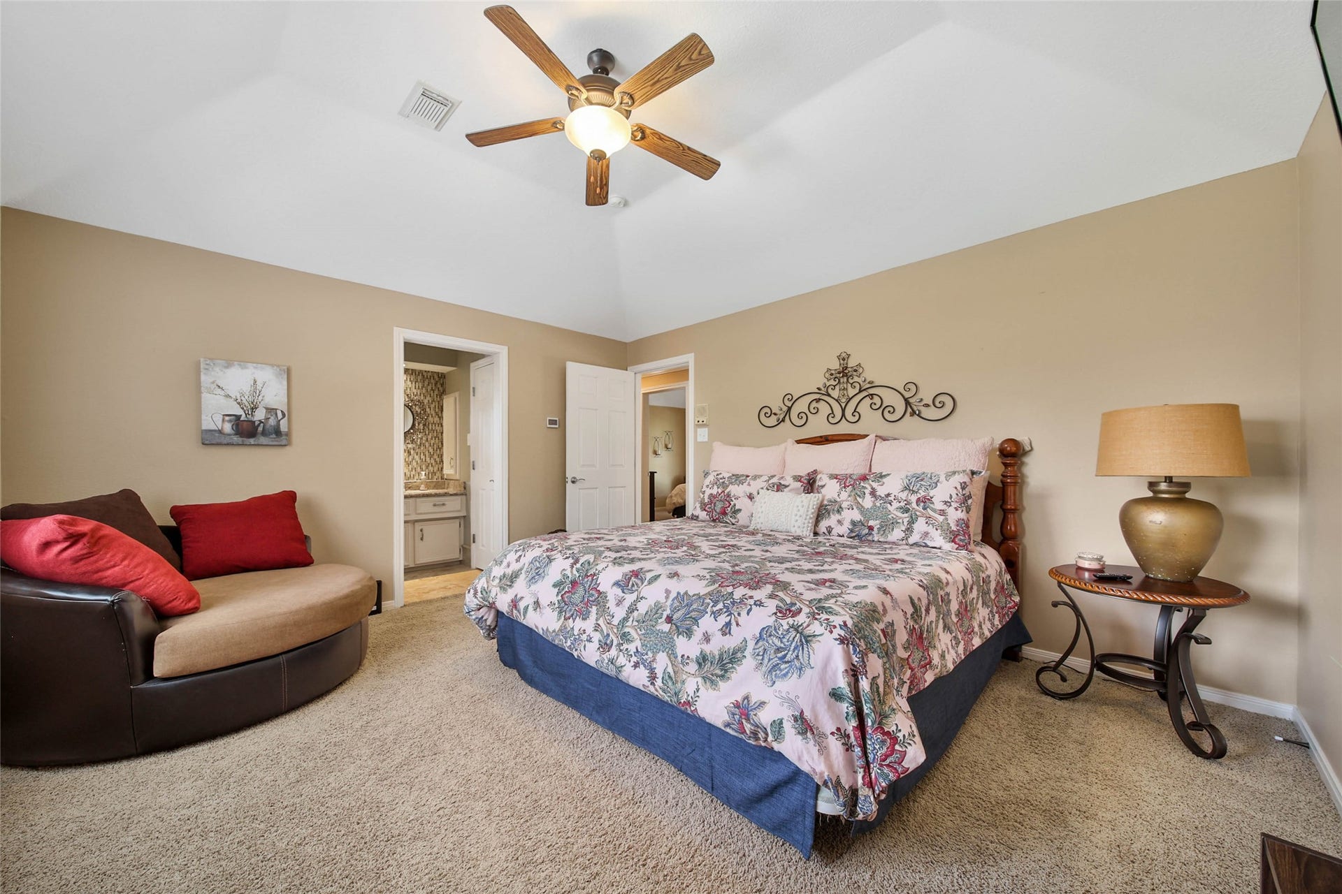 This bedroom features a cozy design with a floral-patterned bed, neutral walls, and carpeted flooring. It includes a ceiling fan, decorative wall art, and access to an en-suite bathroom. A stylish chair and a bedside table with a lamp add comfort and functionality.