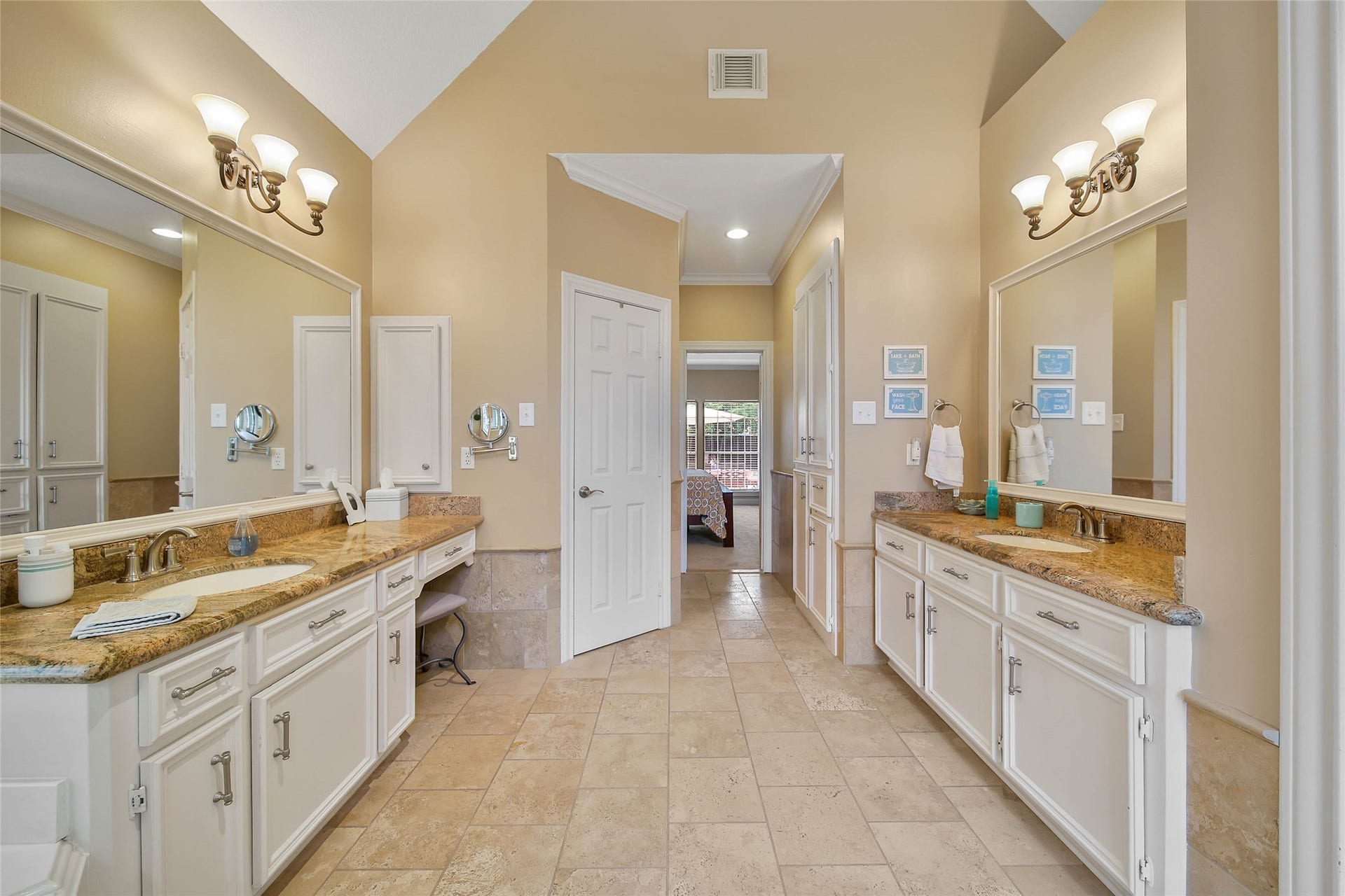 This spacious bathroom features dual vanities with granite countertops, elegant lighting fixtures, and ample cabinet storage. The tile flooring complements the neutral wall tones, creating a warm and inviting atmosphere. A doorway leads to the adjacent bedroom.