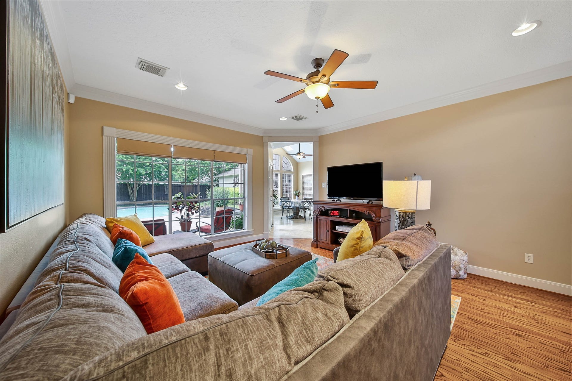 This cozy living room features a large sectional sofa with colorful cushions, a ceiling fan, and a wall-mounted TV. The space is bright with natural light from a sliding glass door that opens to a backyard with a pool. Wood floors and neutral walls create a warm, inviting atmosphere.