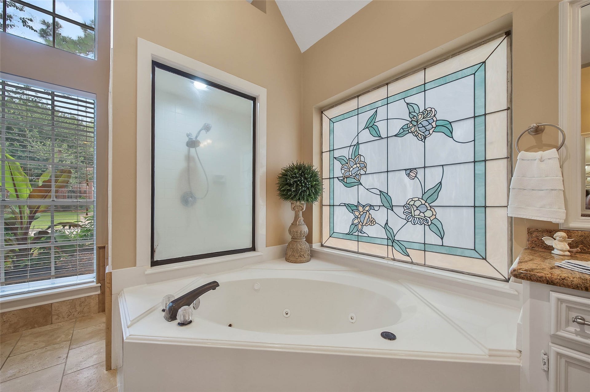 This bathroom features a luxurious corner jacuzzi tub with a stained glass window, providing privacy and style. The space is accented with a frosted glass shower, natural light from large windows, and elegant tile flooring, creating a serene and inviting atmosphere.
