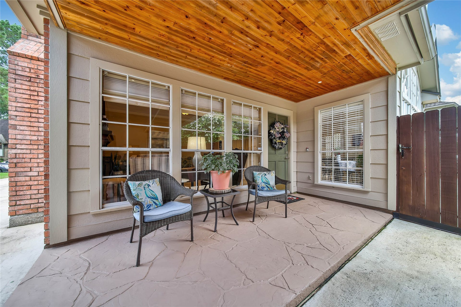 This inviting porch features a warm wooden ceiling, two large windows, and a cozy sitting area with wicker chairs and a small table. It offers a charming entrance with a decorative wreath on the door and a side gate for privacy.