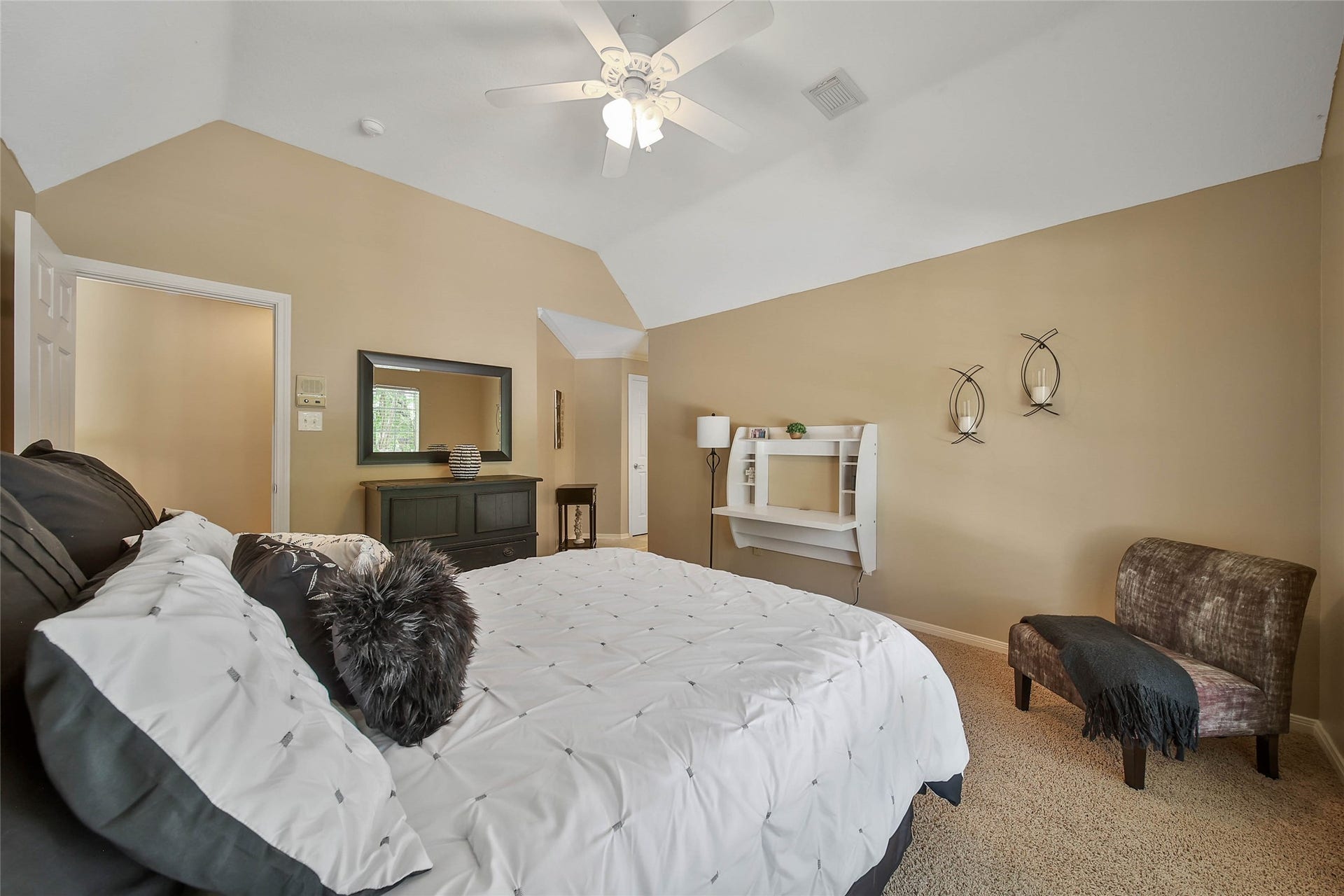 This cozy bedroom features a neutral color palette with a sloped ceiling and ceiling fan. It includes a comfortable bed, decorative wall accents, a small cabinet with a mirror, and a cozy chair, creating a warm and inviting atmosphere.