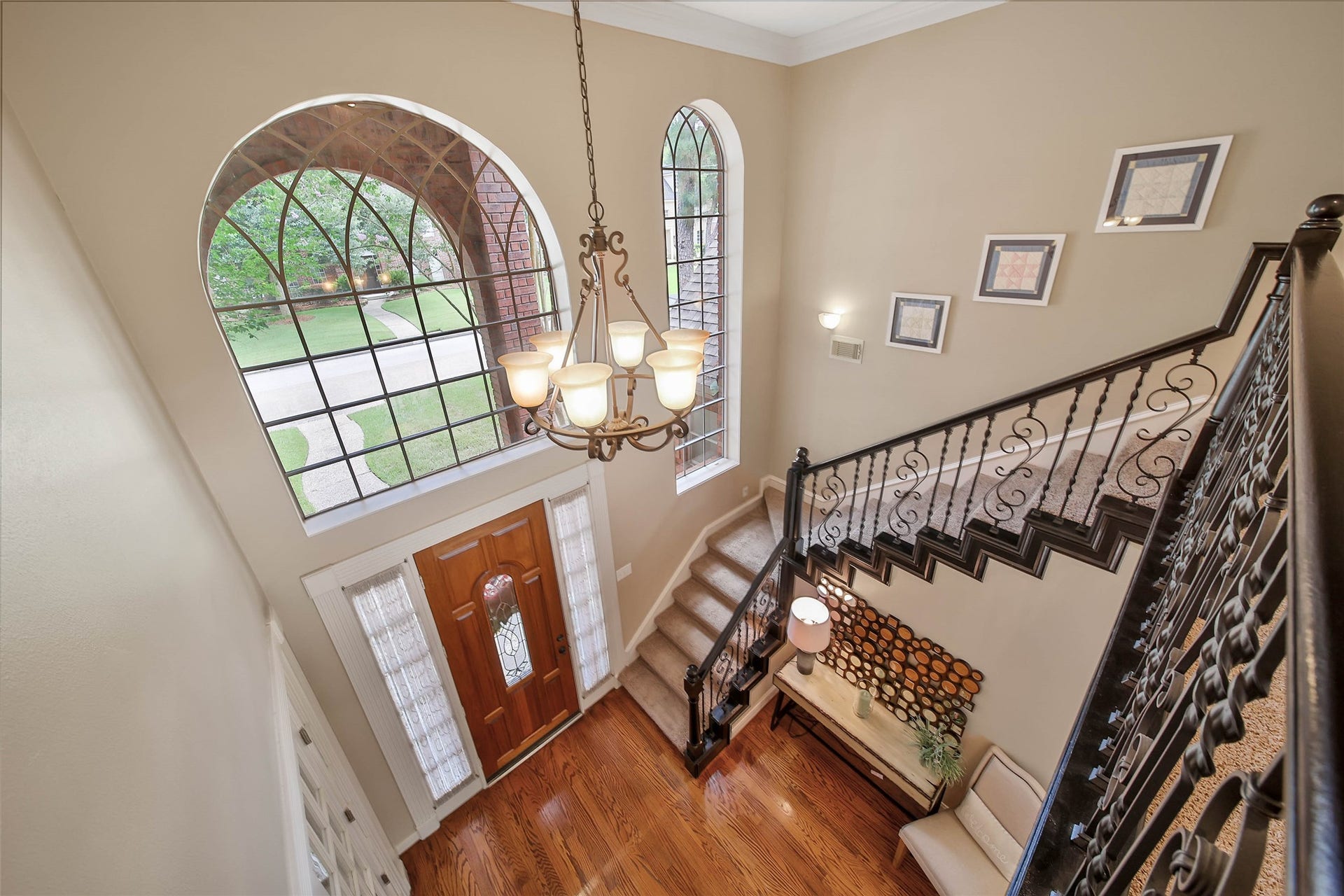 This photo showcases a grand entryway with high ceilings, featuring elegant chandeliers and large arched windows that let in natural light. The staircase has ornate iron railings, and the space is accented with tasteful decor and warm wood flooring.