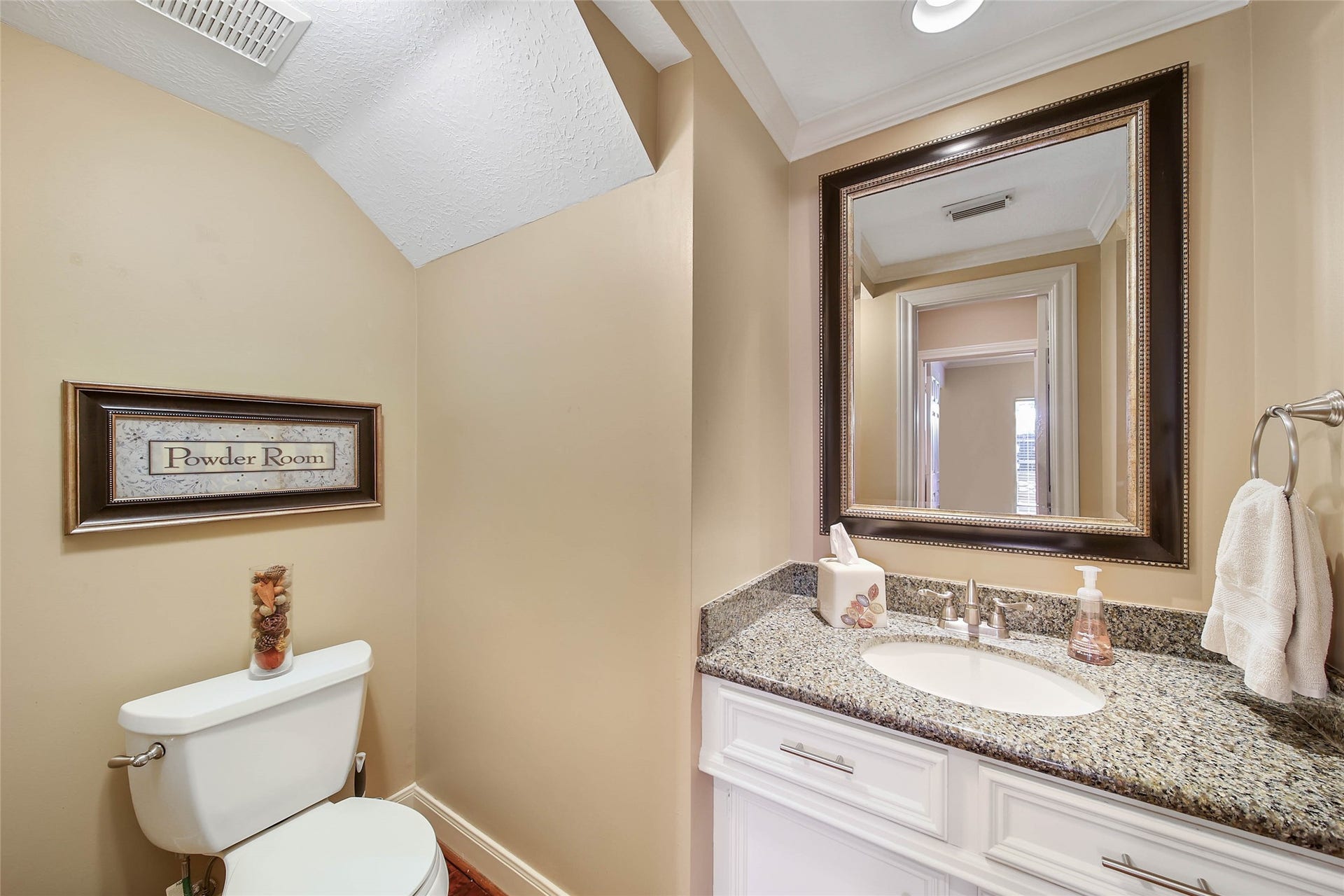 This powder room features a sleek granite countertop, a large framed mirror, and warm beige walls. It includes a toilet with decorative accents and a towel ring, creating a cozy and inviting space.