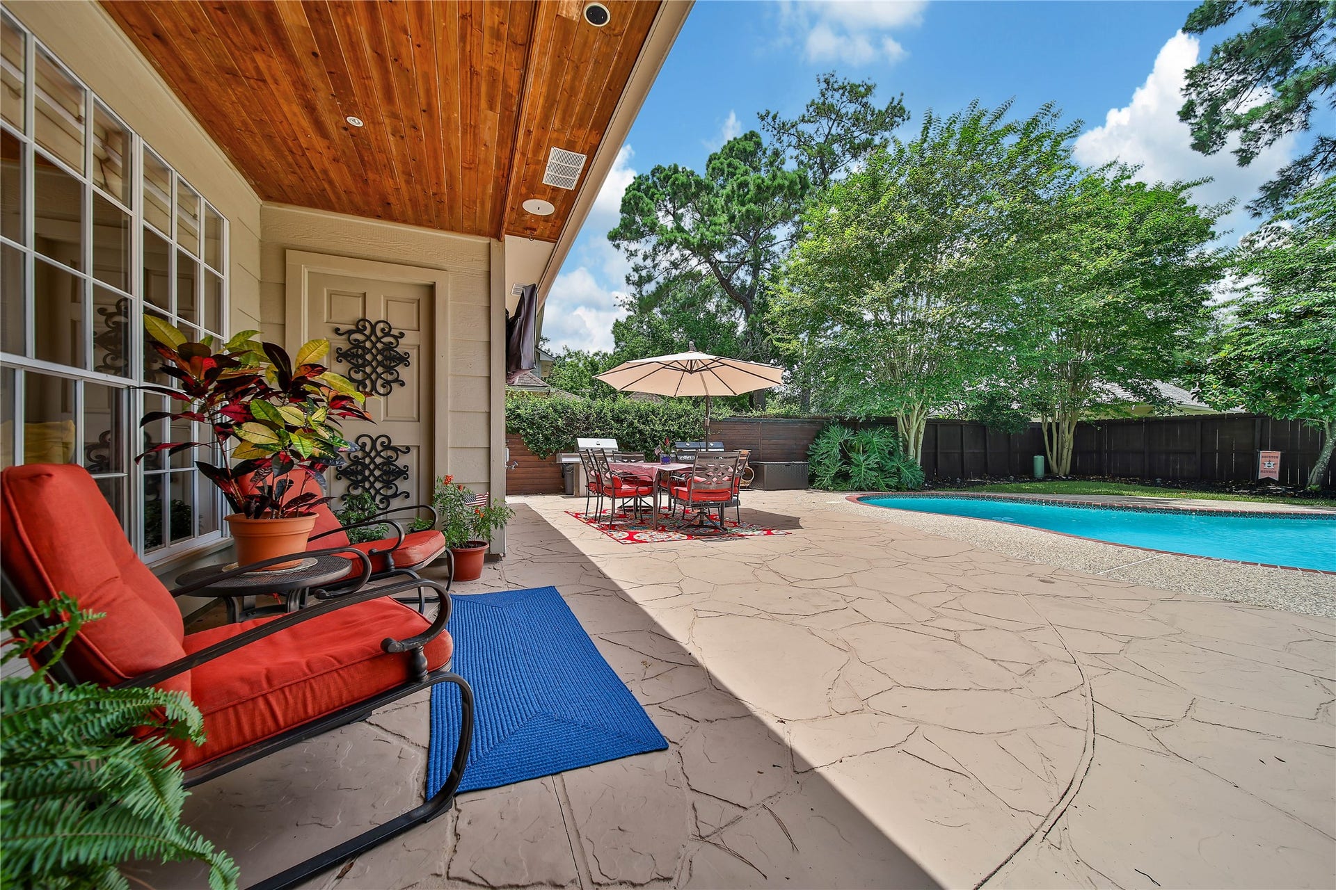 This backyard oasis features a spacious patio with cozy seating, vibrant plants, and a dining area under an umbrella. It overlooks a refreshing pool, surrounded by lush greenery for privacy and tranquility. Designed for gathering inside and out, with multiple living spaces and an outdoor setting that becomes part of the home.