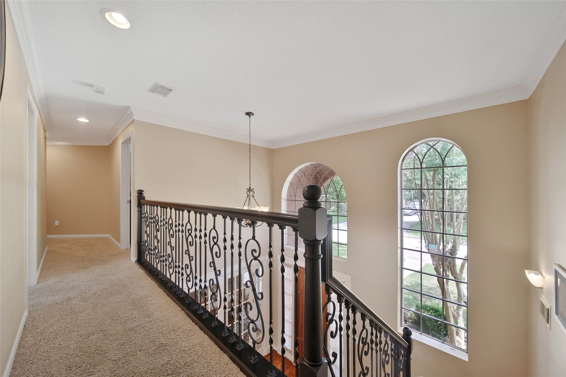 This photo shows an elegant upstairs hallway with a wrought-iron railing overlooking large arched windows that provide natural light. The space is carpeted and features soft, neutral walls, creating a warm and inviting atmosphere.