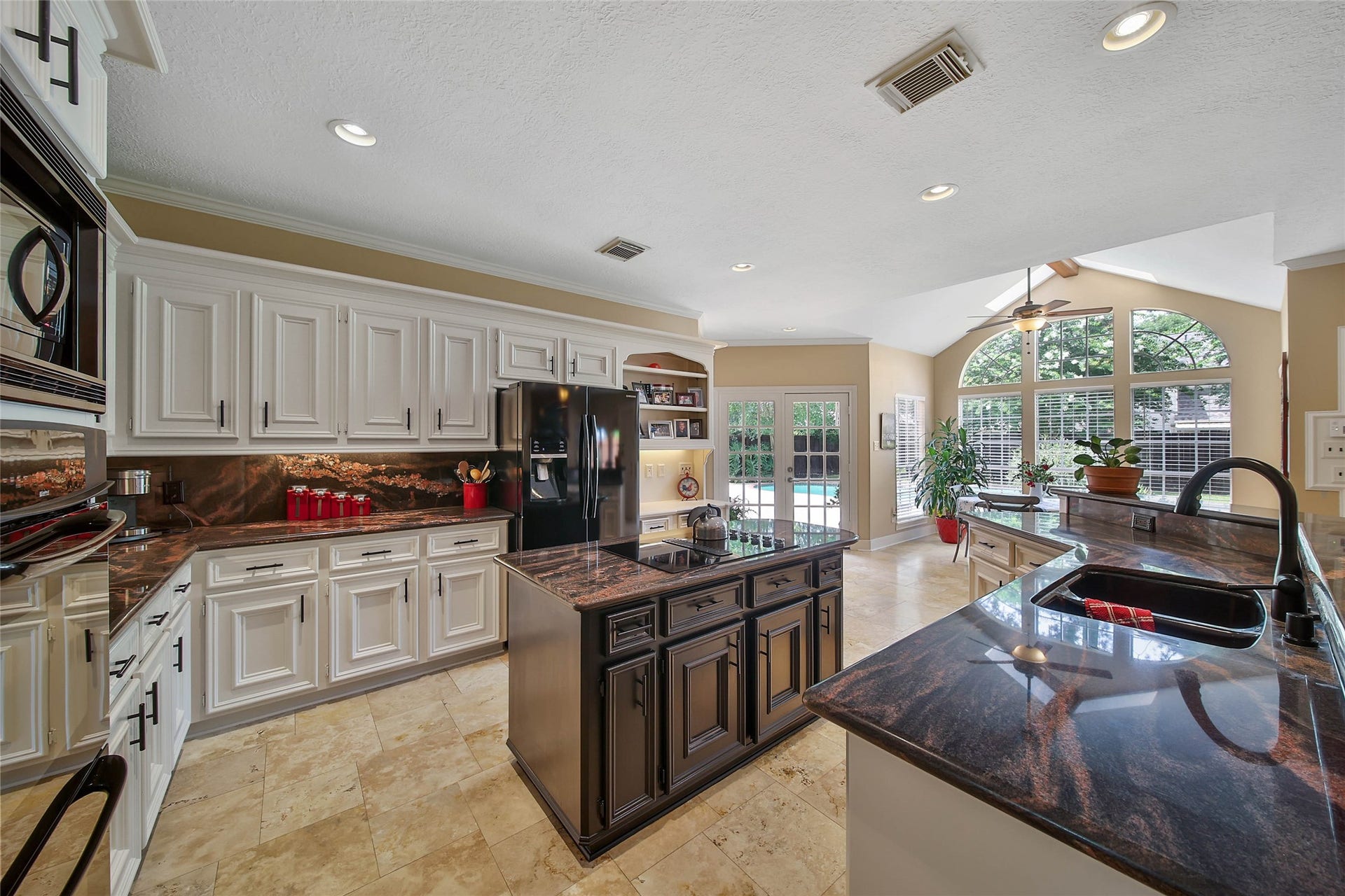 This spacious kitchen features elegant white cabinetry, dark countertops, and a central island. It opens to a bright living area with large windows and views of the backyard pool, creating a perfect blend of style and functionality.