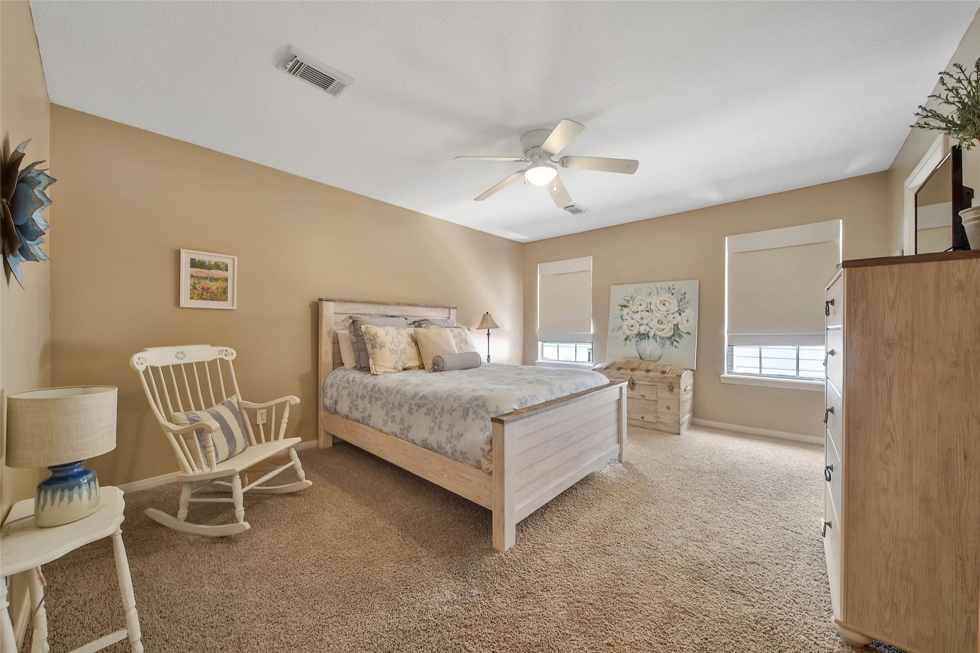 Spacious bedroom with neutral tones featuring a comfortable bed, a ceiling fan, and ample natural light from two windows. It includes cozy furnishings like a rocking chair, dresser, and decorative accents, creating a warm and inviting atmosphere.