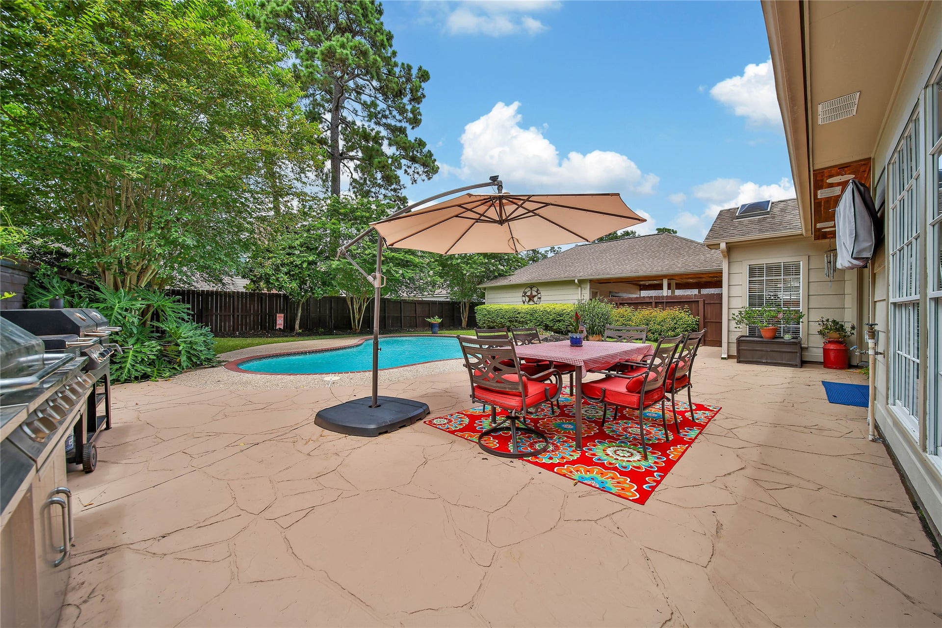 This backyard features a spacious patio with a dining set under a large umbrella, a built-in grill, and a colorful rug. It overlooks a well-maintained pool surrounded by lush greenery, offering a perfect space for outdoor entertaining and relaxation.