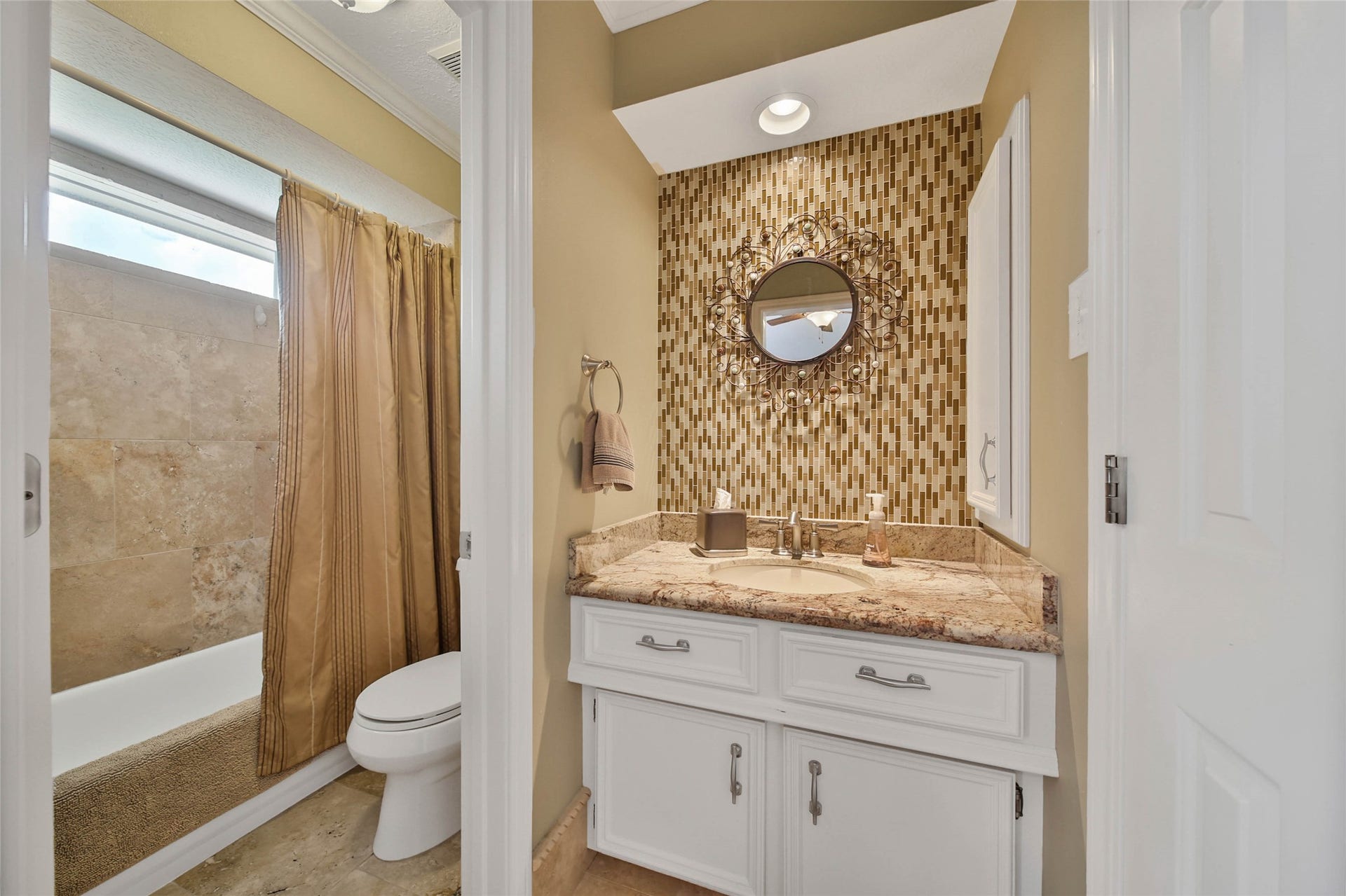 This photo features a stylish bathroom with a tiled shower and a classic white bathtub. The vanity area boasts a granite countertop and an ornate circular mirror set against a mosaic tile accent wall, adding a touch of elegance.