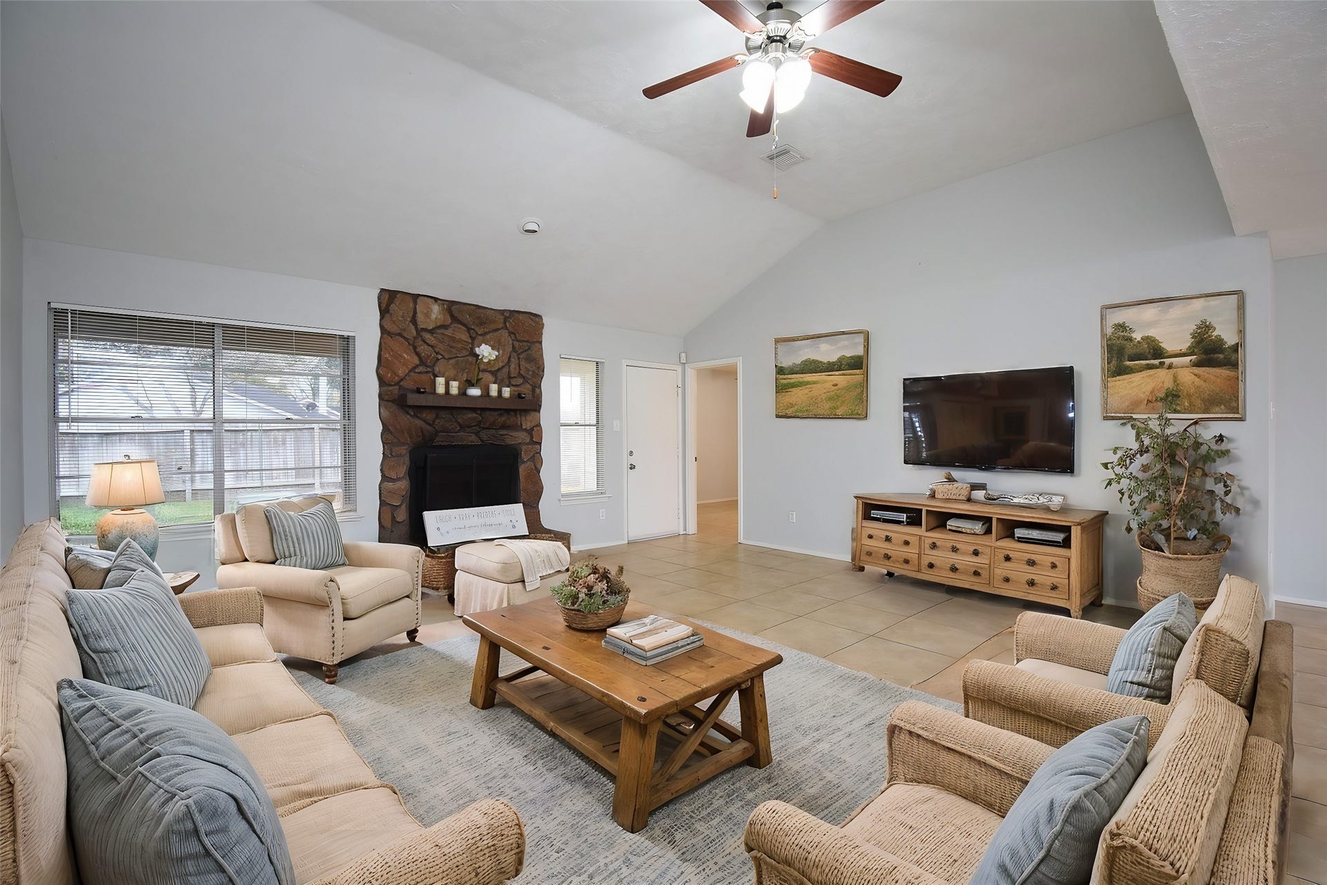 16806 Red Gully Drive, Sugar Land, TX 77498 virtual staged : This spacious living room features a vaulted ceiling with a ceiling fan, a cozy stone fireplace, and large windows for natural light. The neutral tones and tile flooring create a warm, inviting atmosphere.