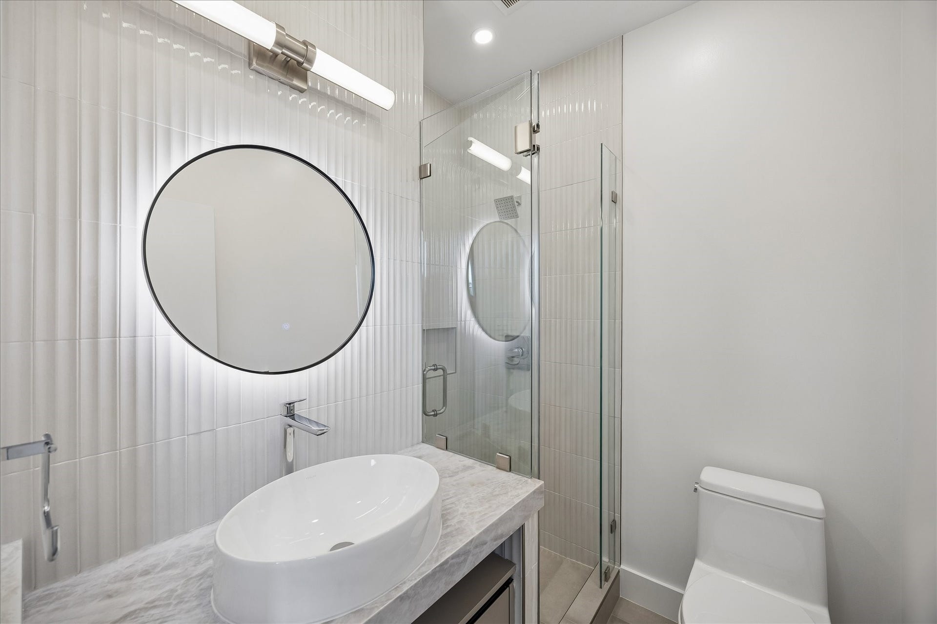 Modern bathroom with glass-enclosed shower, stylish round mirror with backlighting, and elegant vessel sink on a marble countertop.