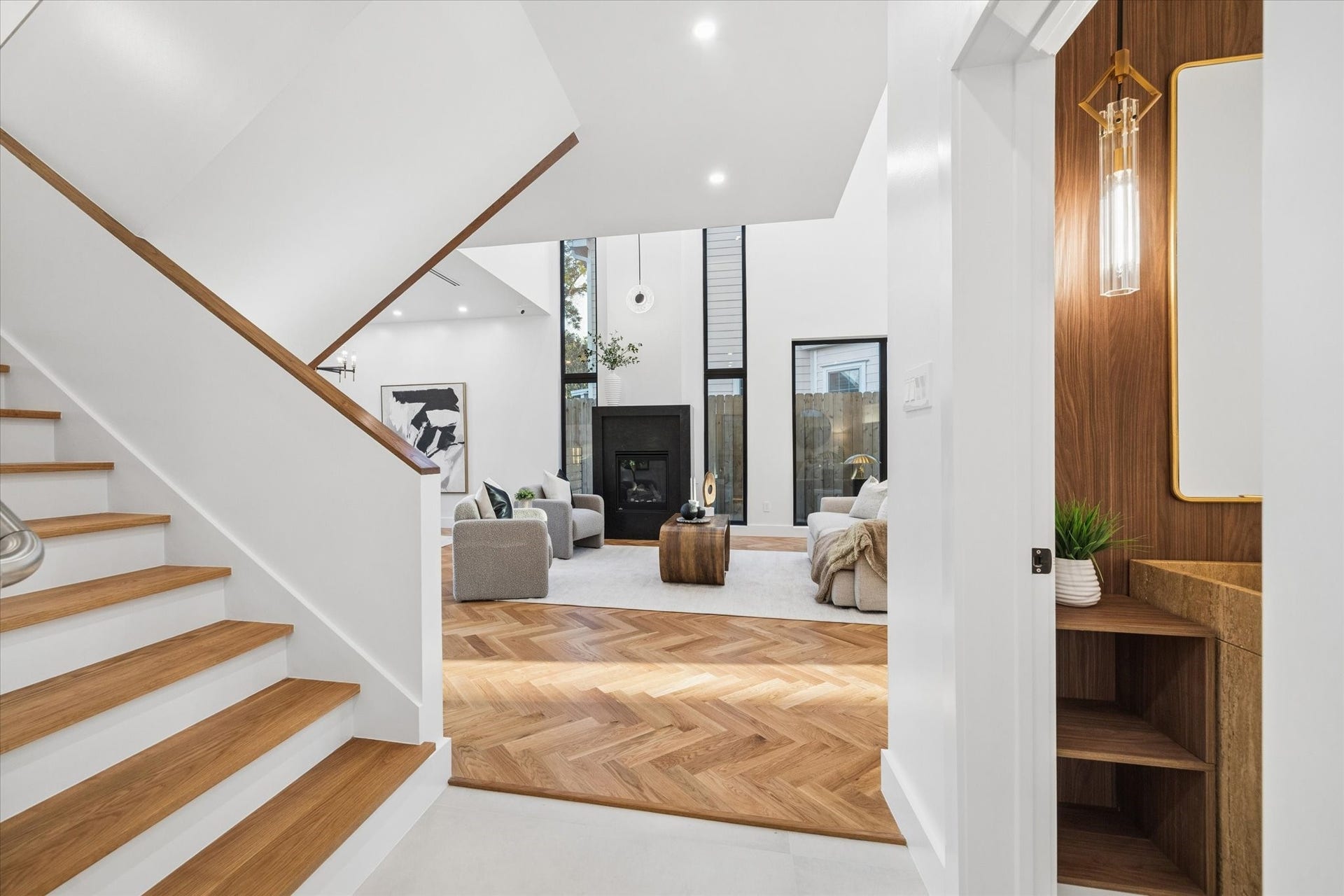 Discover a stunning open-concept living area featuring sleek wood flooring, a cozy fireplace, and abundant natural light from large windows, all viewed from a modern staircase.