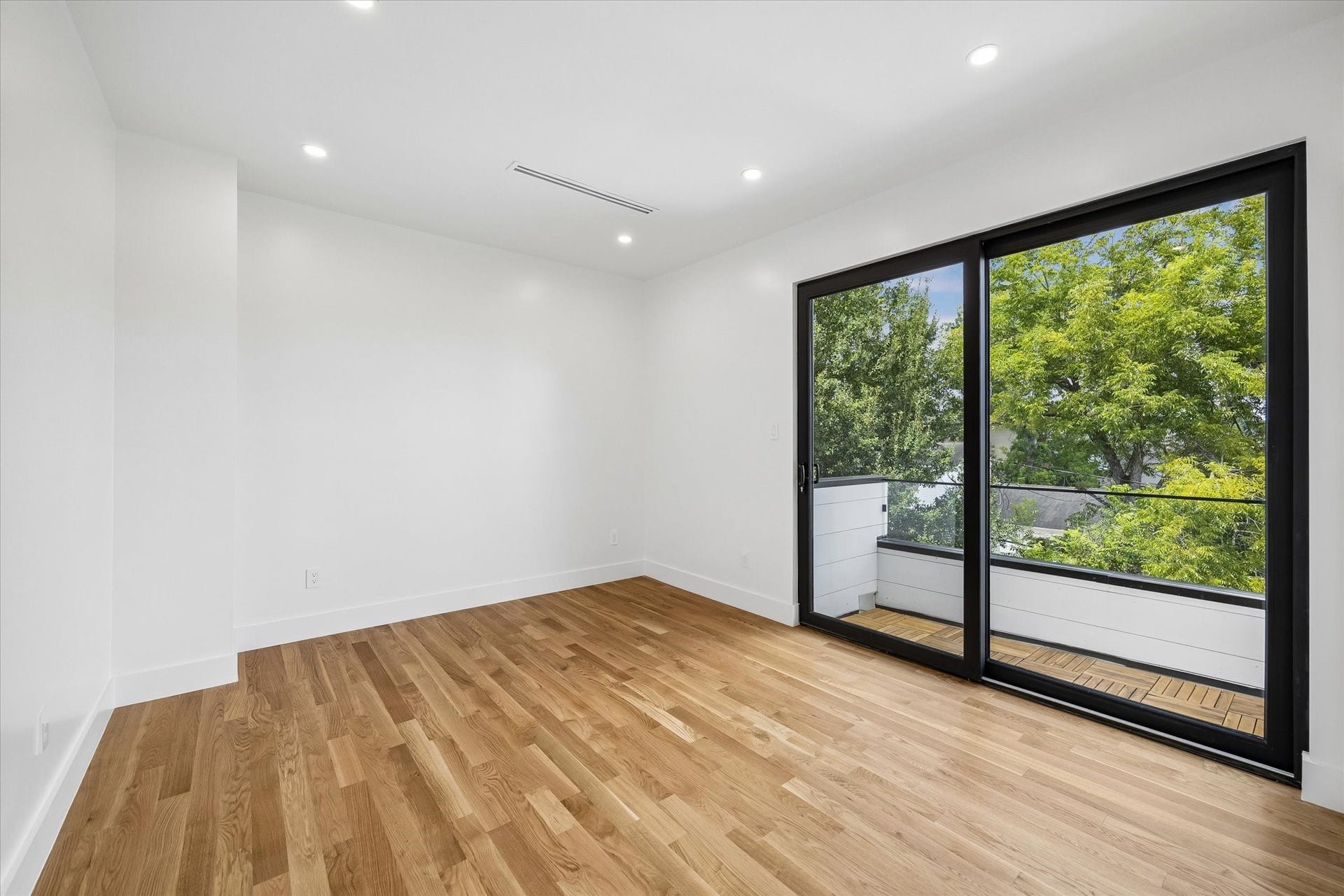 Bright and spacious room with hardwood floors, large sliding glass doors, and a private balcony offering a view of greenery.