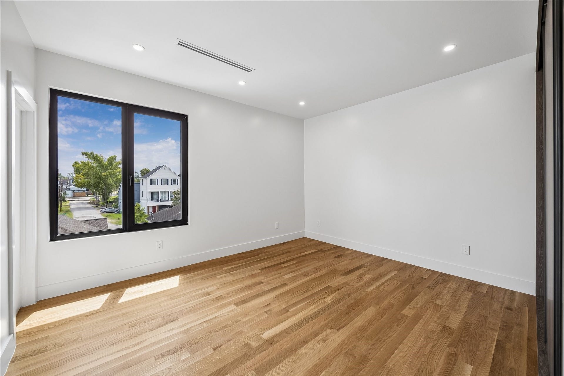 Spacious, sunlit room with hardwood floors and a large window offering neighborhood views.