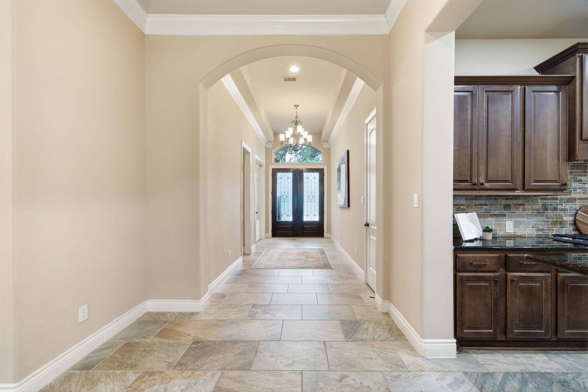 4603 Piper Shadow Lane, Katy, TX 77494 Elegant entryway featuring a grand archway, stylish chandelier, and gleaming tile floors, leading to a welcoming front door with frosted glass panels. Adjacent kitchen boasts rich wooden cabinetry and stone backsplash.